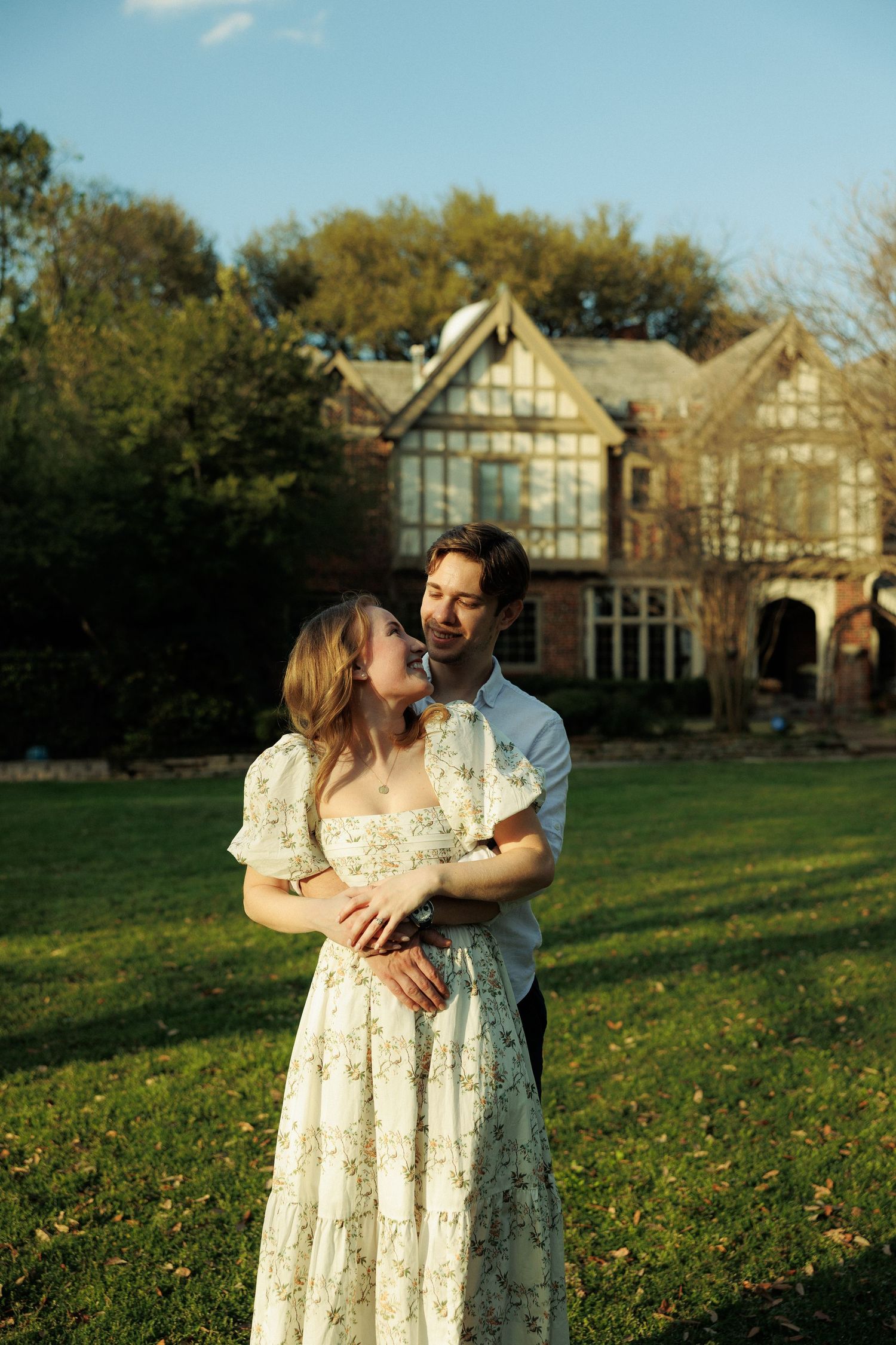 A couple embraces on a sunny lawn with Tudor-style buildings visible in the background during a romantic photoshoot.
