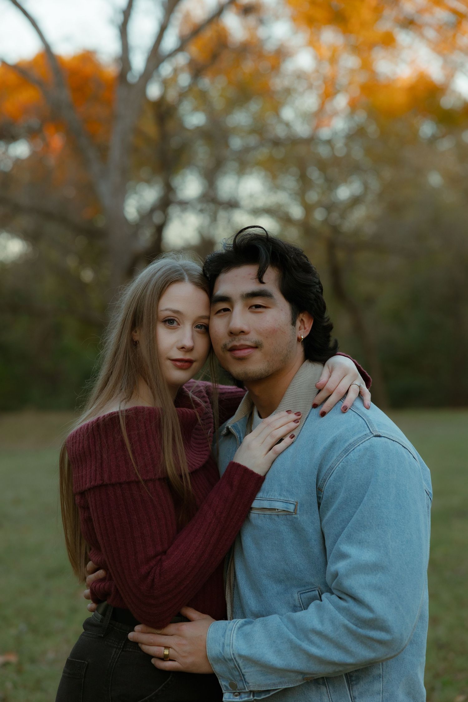 A couple embraces outdoors during autumn with orange foliage in the background.