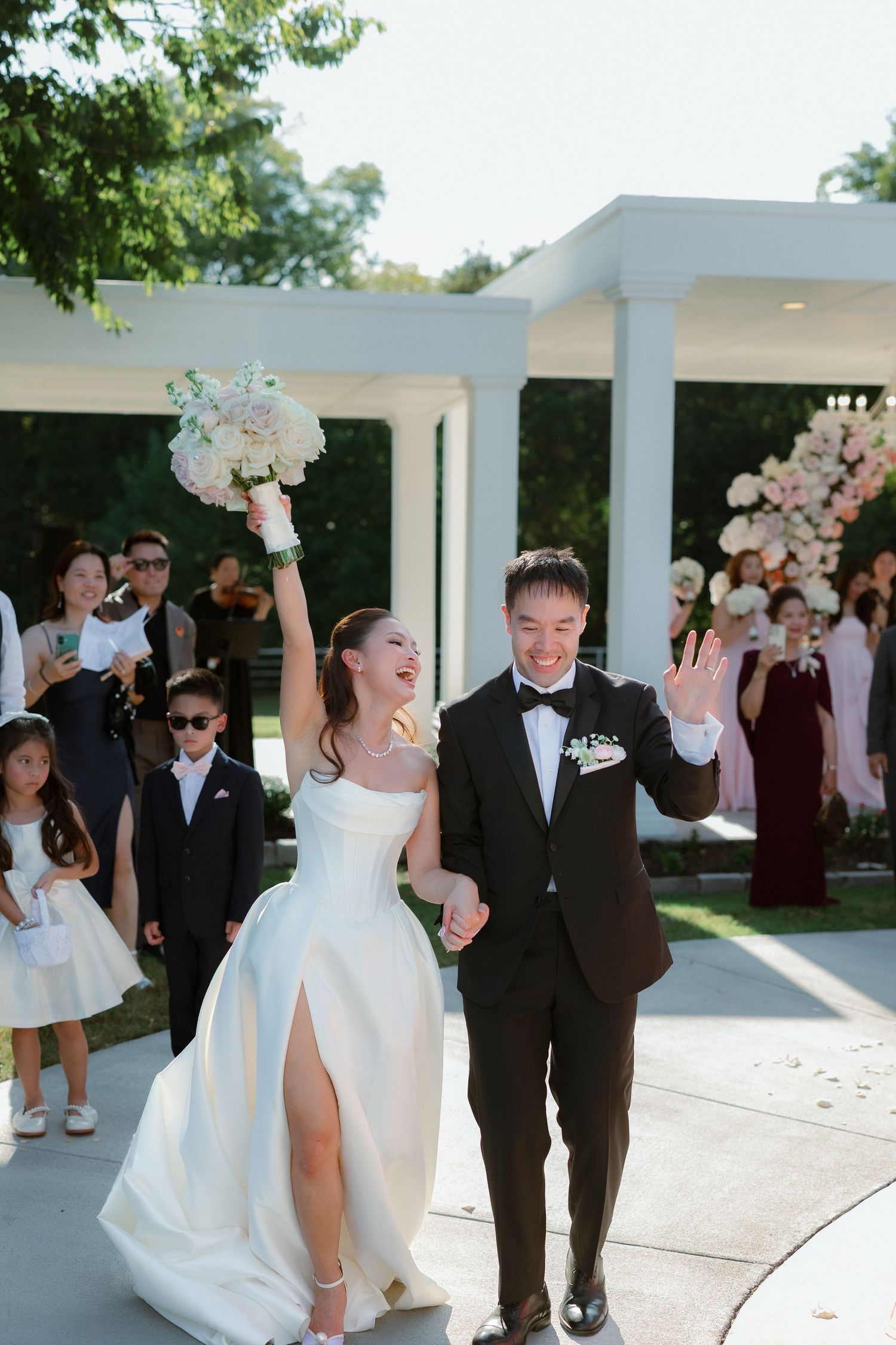A wedding party celebrates under white columns outdoors with bridesmaids in burgundy dresses and groomsmen in black tuxedos.