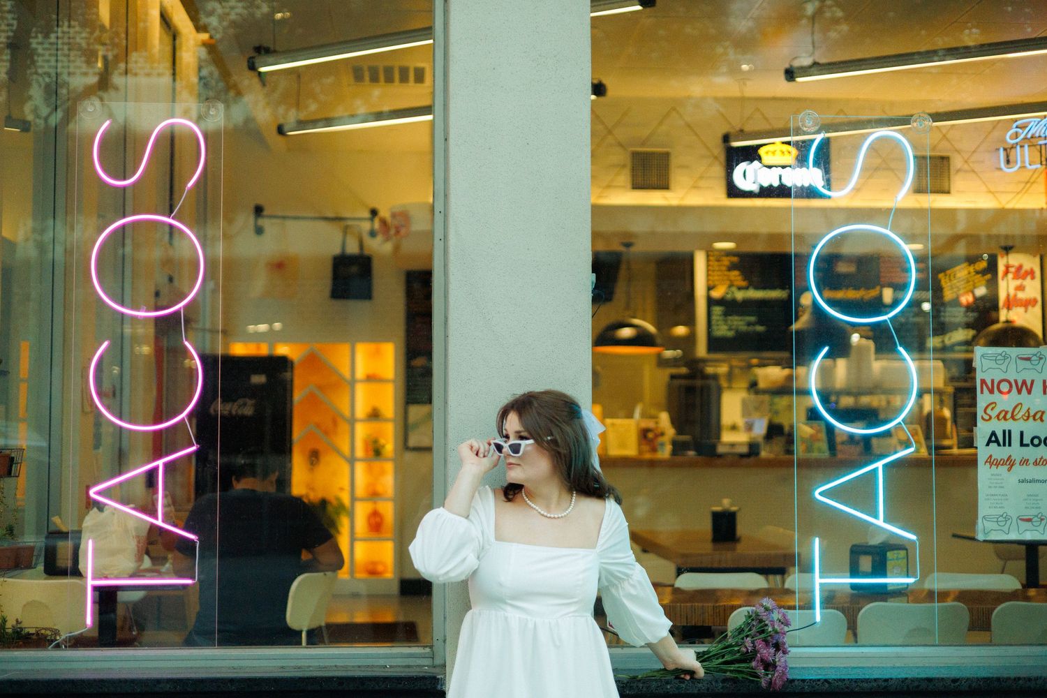 Bride in white dress standing by storefront window with illuminated TACOS signage reflected in glass.