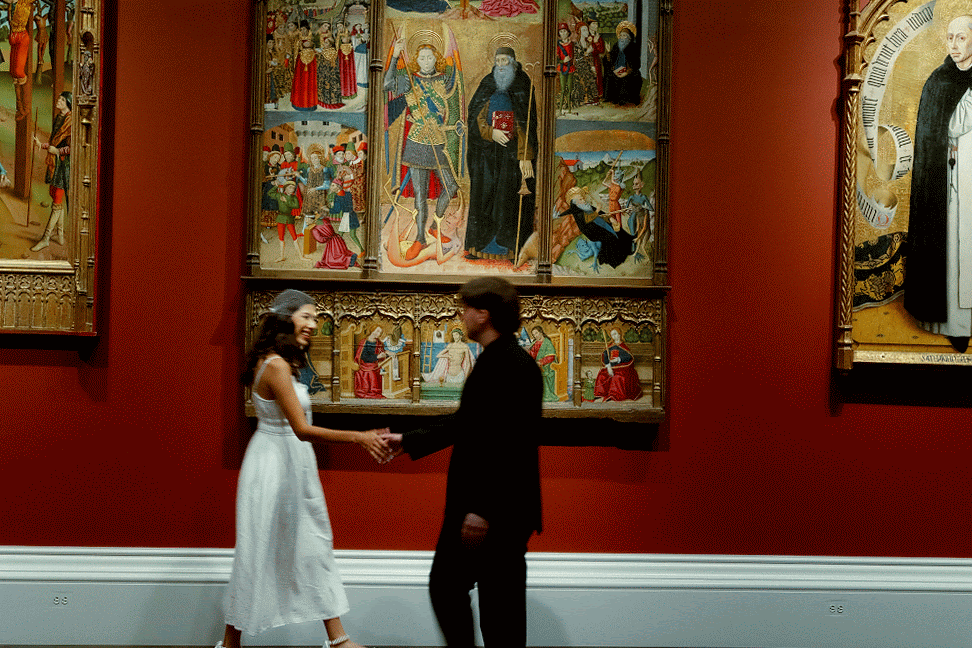 Art gallery visitors admire historic paintings displayed on deep red walls in a museum exhibition space.