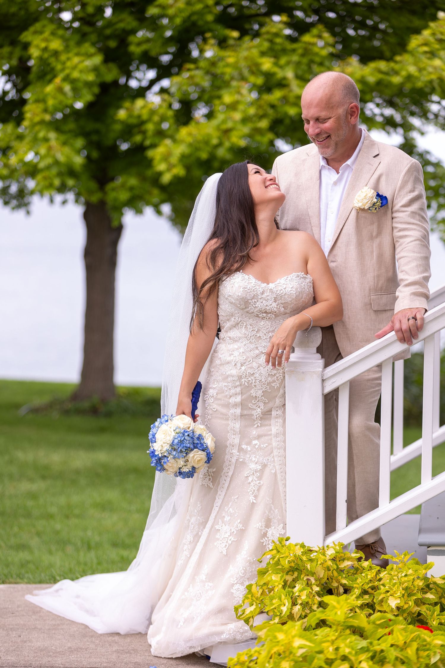 A couple in wedding attire share a romantic moment on white stairs with blue and yellow flowers in a garden setting.