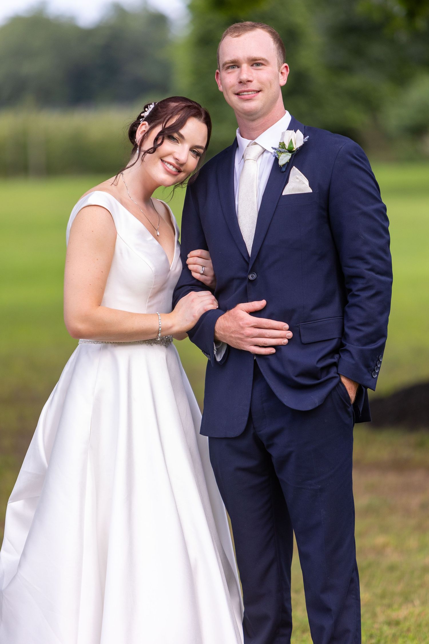 A couple poses outdoors on their wedding day wearing a white gown and navy suit with floral boutonniere.