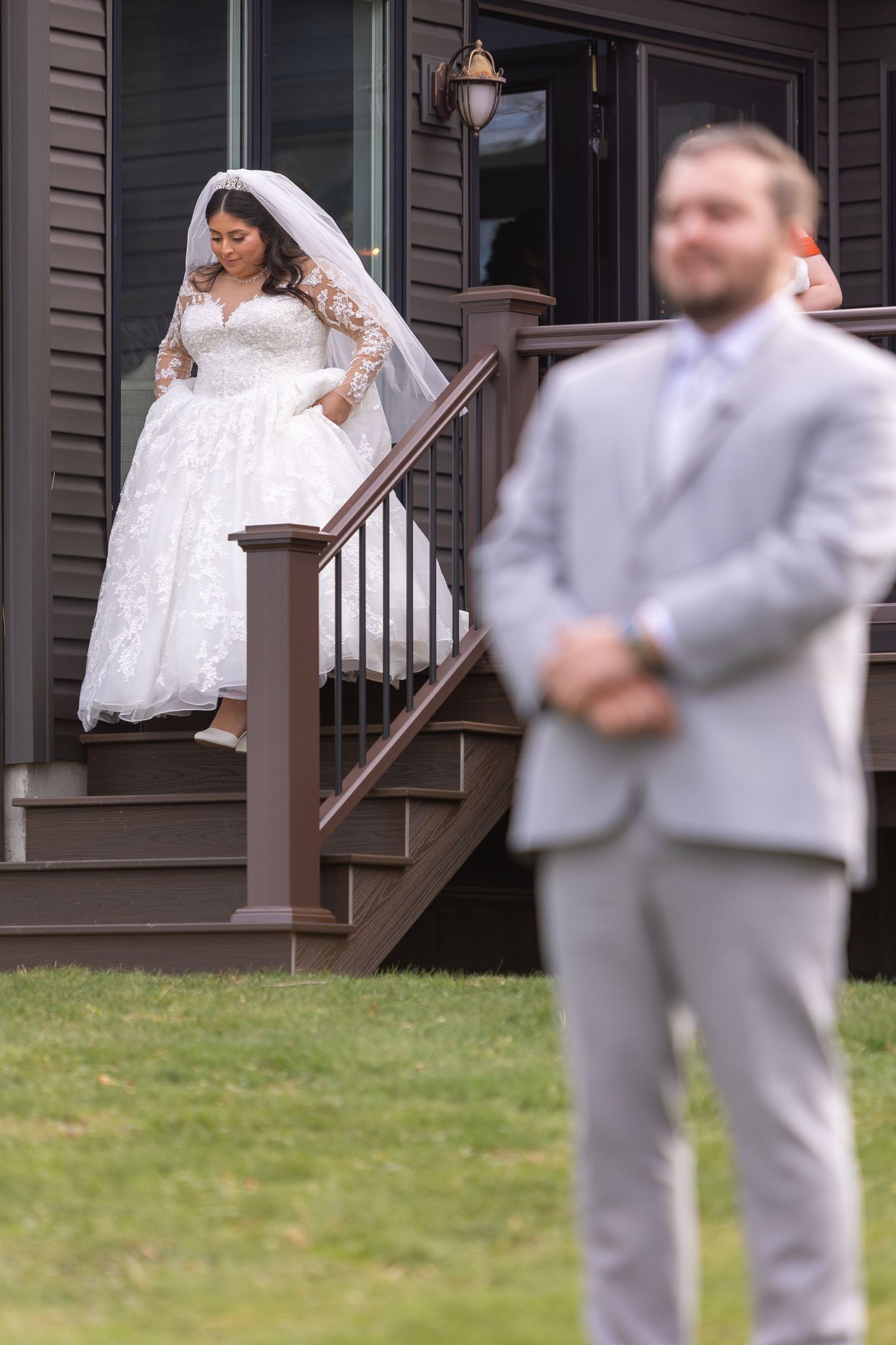 A bride in a lace wedding dress stands on wooden stairs while a groom in a light gray suit waits in the foreground.