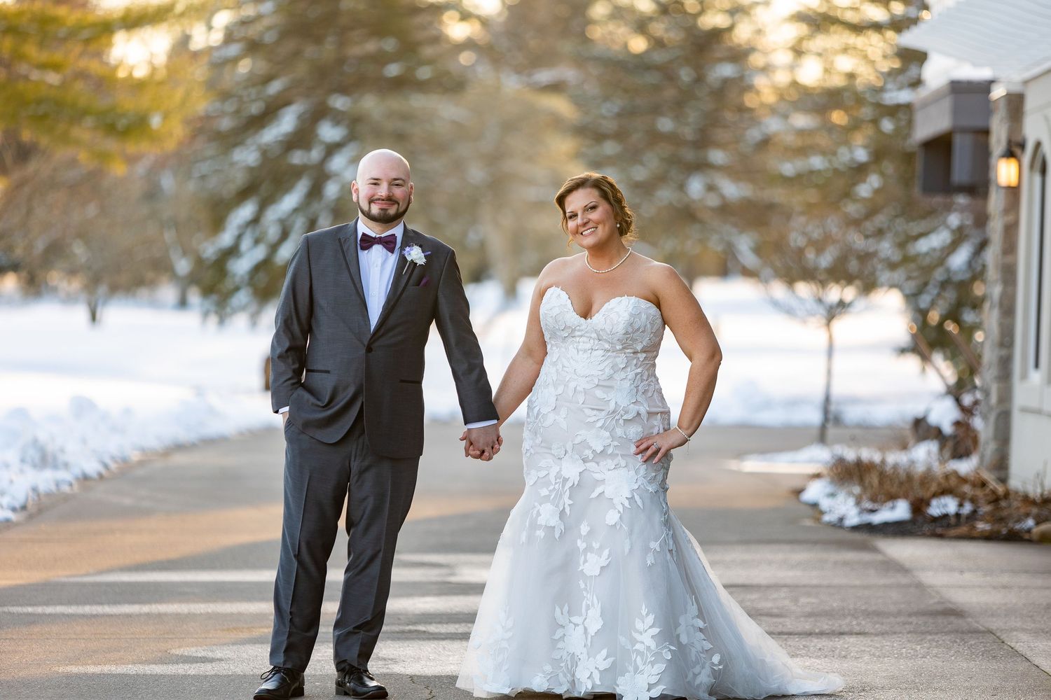 A couple holds hands while walking outdoors on a snowy winter day, with the bride wearing a lace gown and groom in tuxedo.