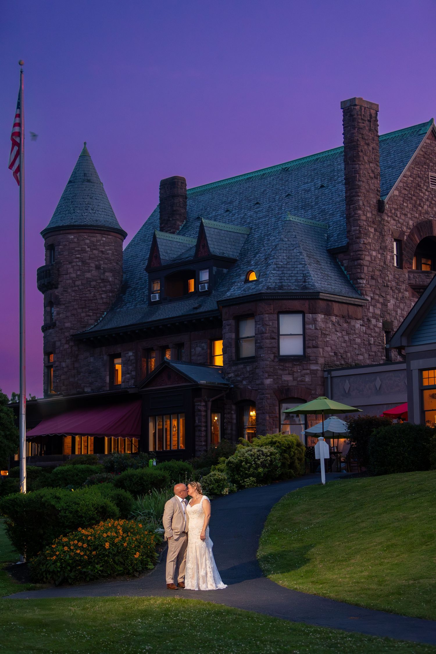 A couple stands on a curved pathway in front of an illuminated stone castle-like building at dusk with a purple sky.