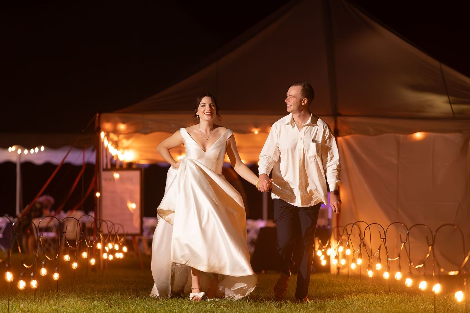 People in matching white outfits dancing outdoors at night under warm glowing string lights at a celebration.