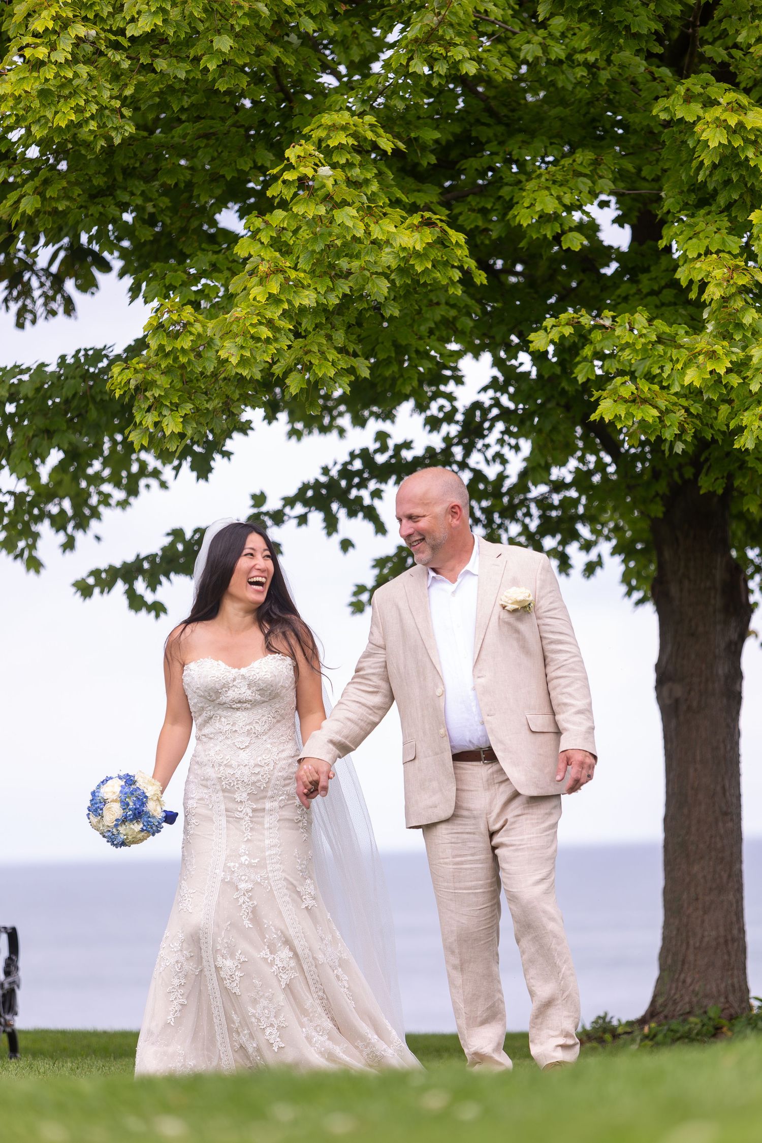 A couple walks on a grassy hill together on their wedding day, with lush green trees in the background.