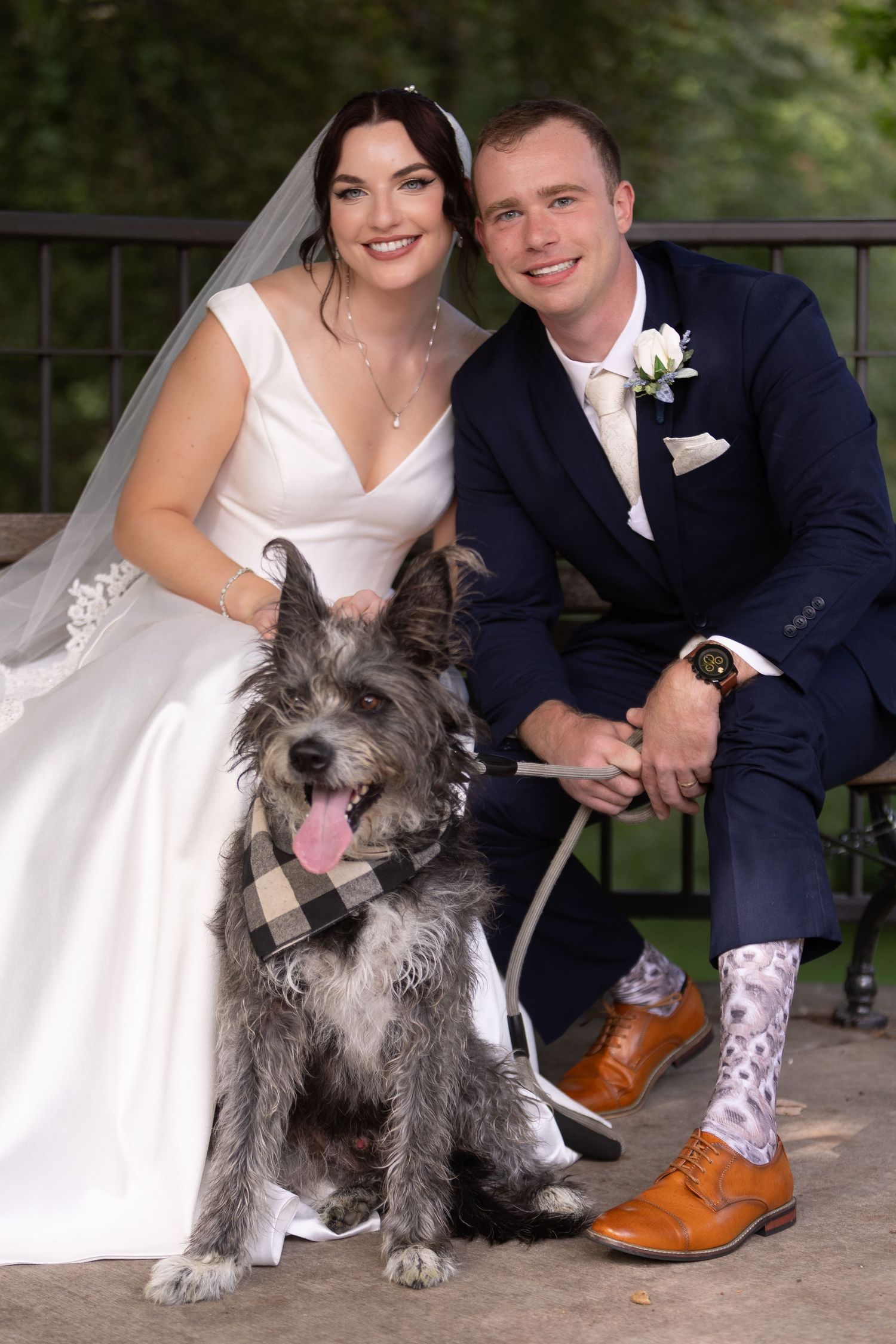 A wedding couple poses with their scruffy grey dog wearing a plaid bowtie on a porch.