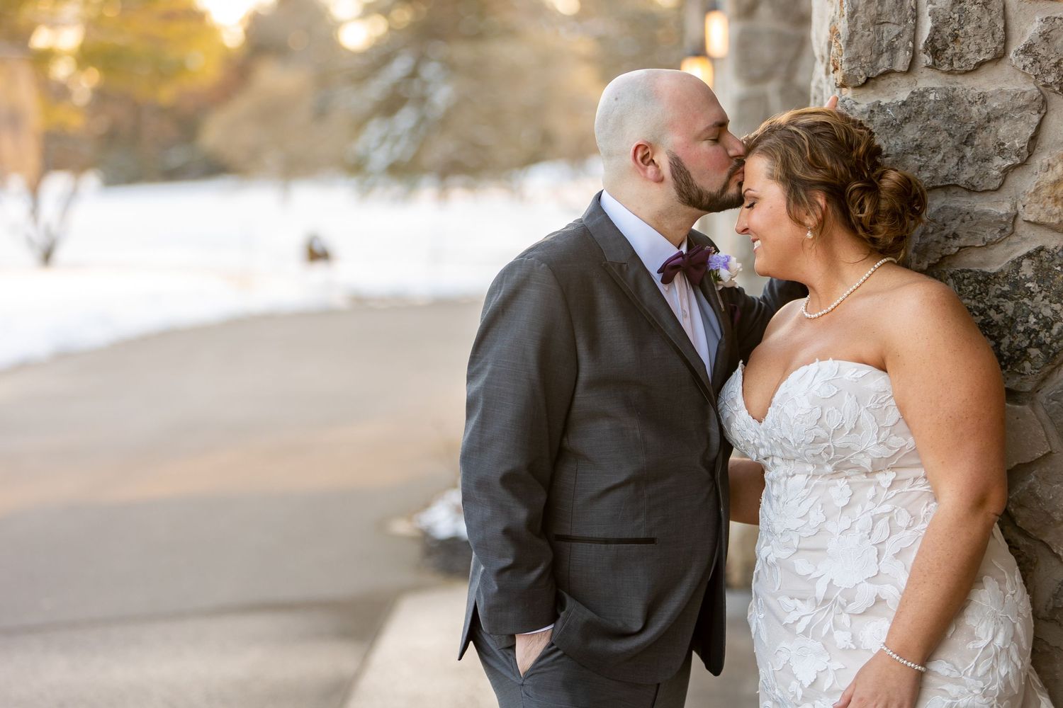 A newlywed couple shares an intimate moment against a stone wall with a snowy winter landscape in the background.