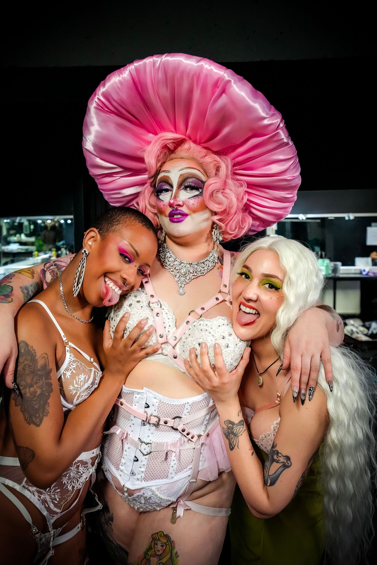 Three performers in pink and white outfits pose together playfully in a series of glamorous backstage photos.