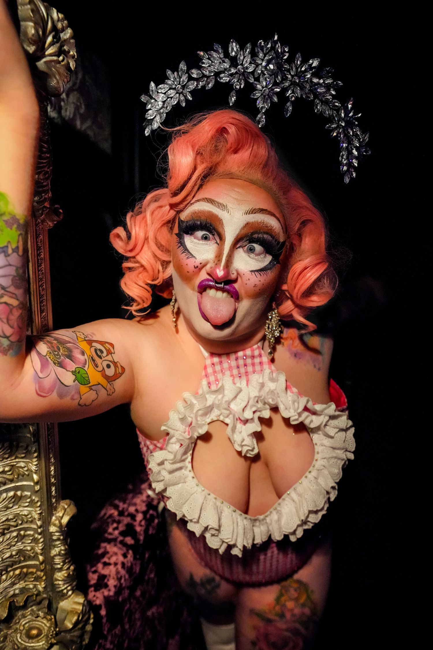 Drag performer with pink hair, silver crown, dramatic makeup, ruffled costume, and colorful tattoos poses playfully.