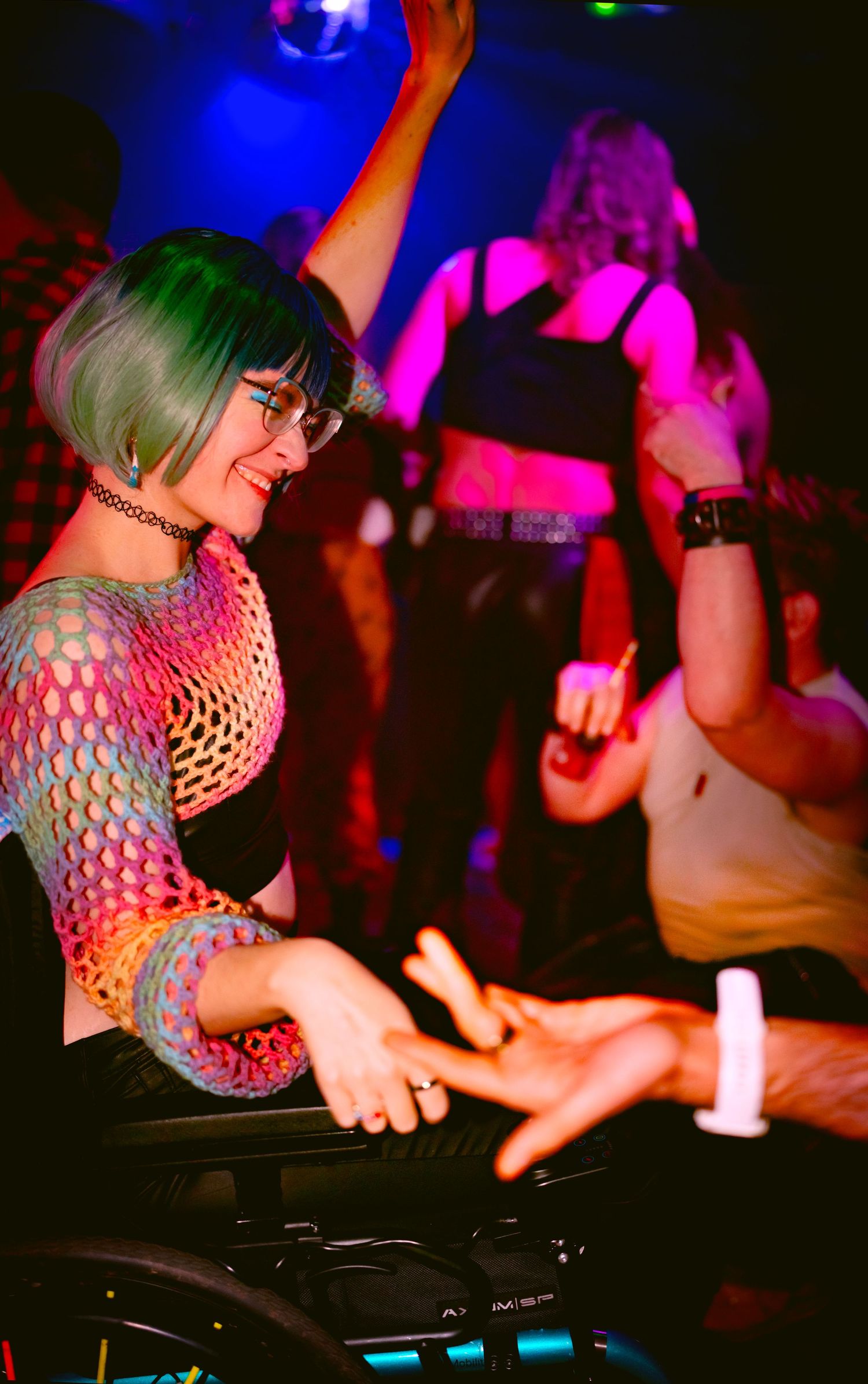 A young woman with green hair in a mesh top. Queer in a wheelchair in a nightclub, smiling while people dance behind her.