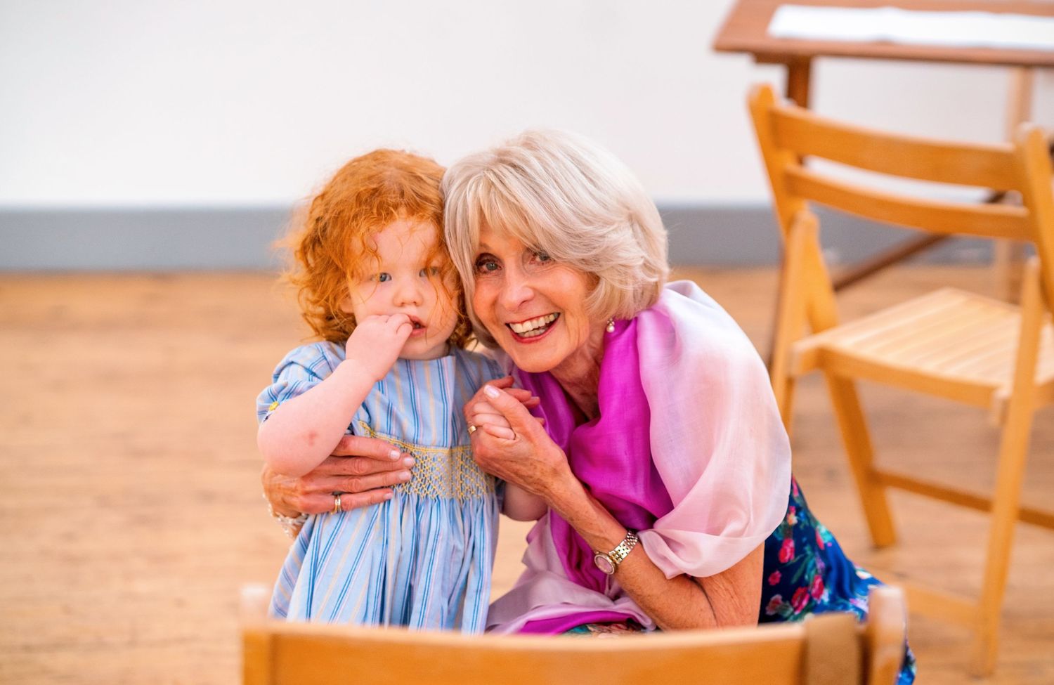 Senior and child sharing a joyful moment together while sitting on wooden chairs in a bright sunny room.