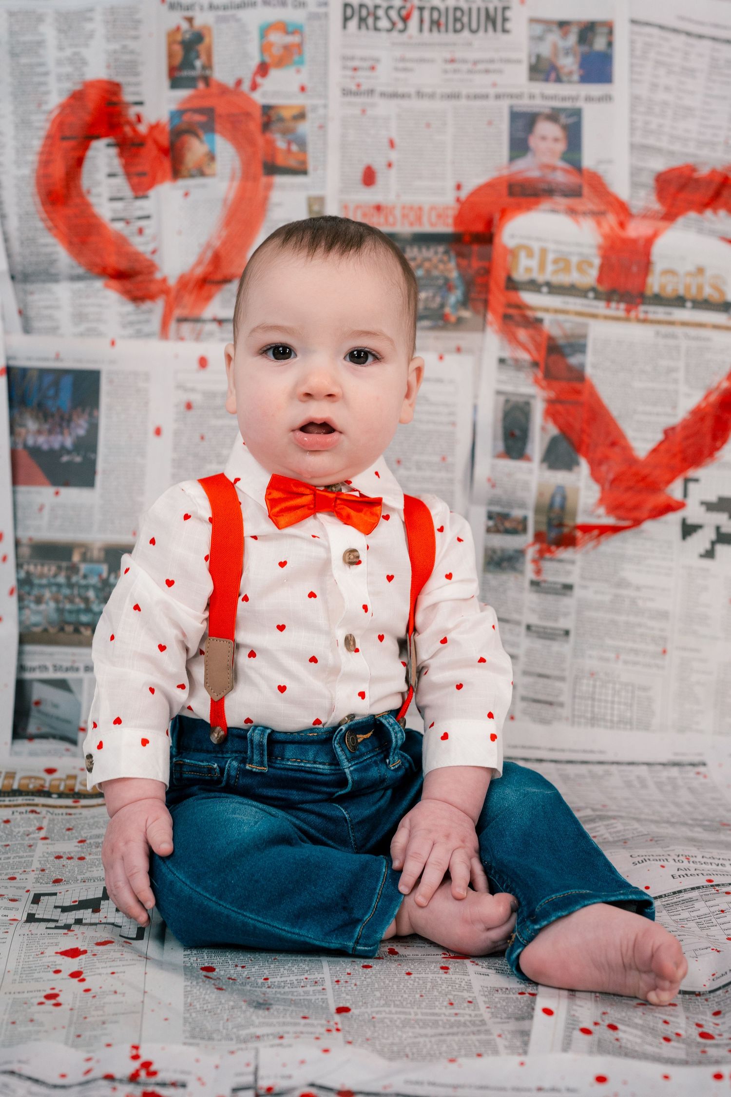 A baby in suspenders and bow tie sits on newspaper backdrop with red heart decorations.