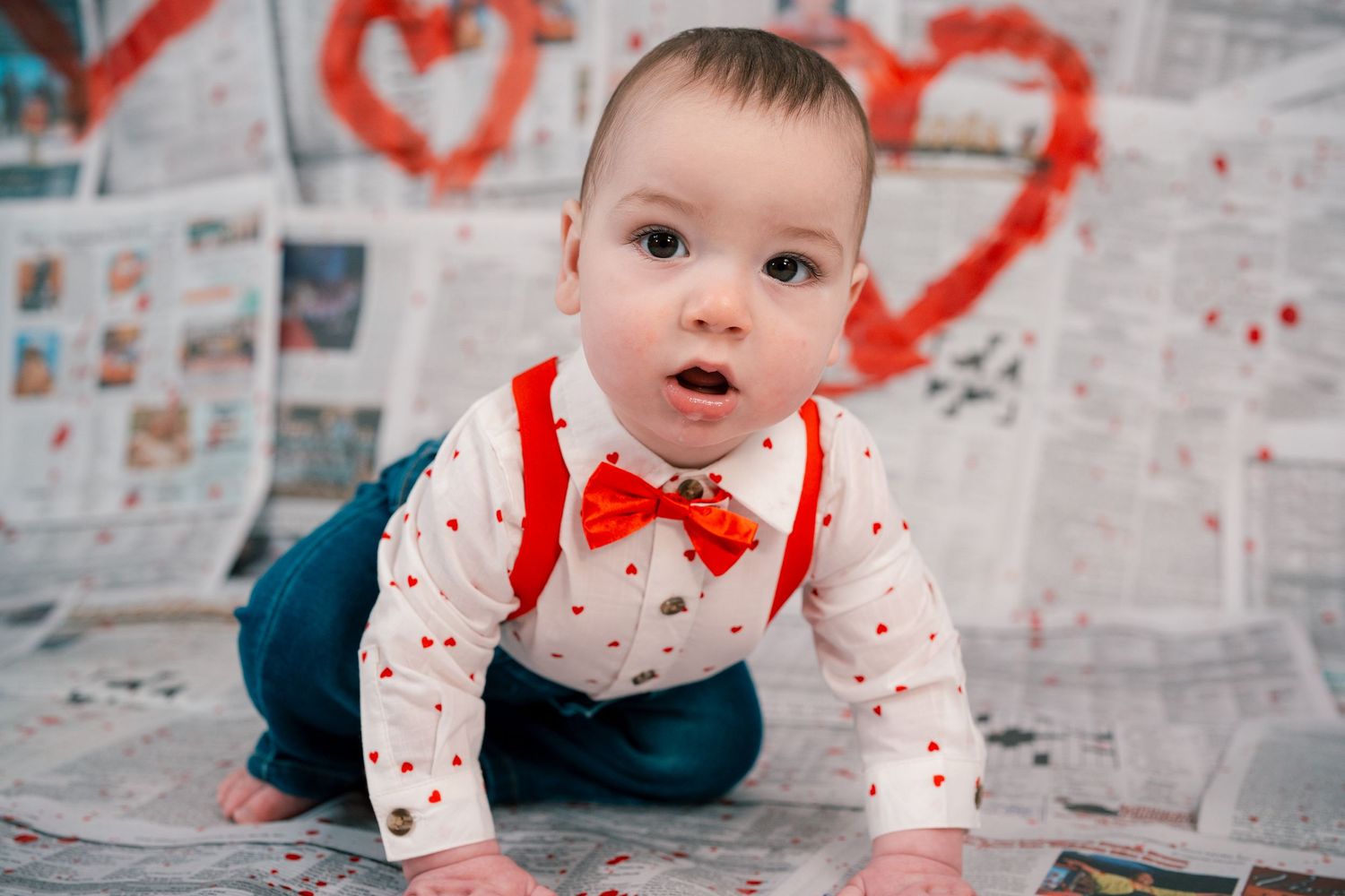 Baby in white shirt with red bow tie crawls on newspaper with painted red hearts in background.