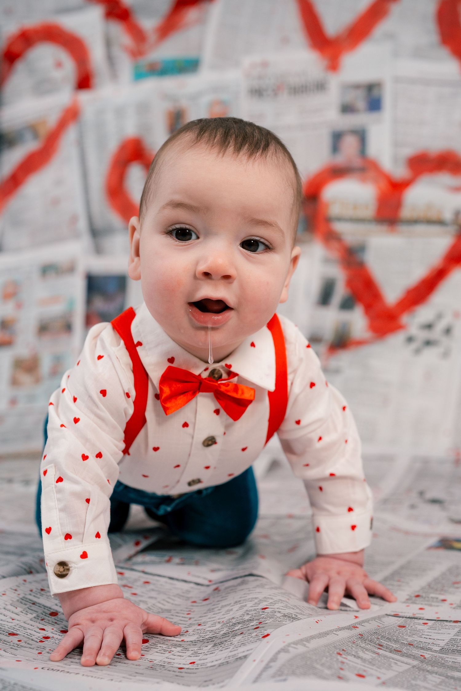 A baby in a heart-patterned outfit with red bow tie crawls on newspaper-covered floor with heart decorations.
