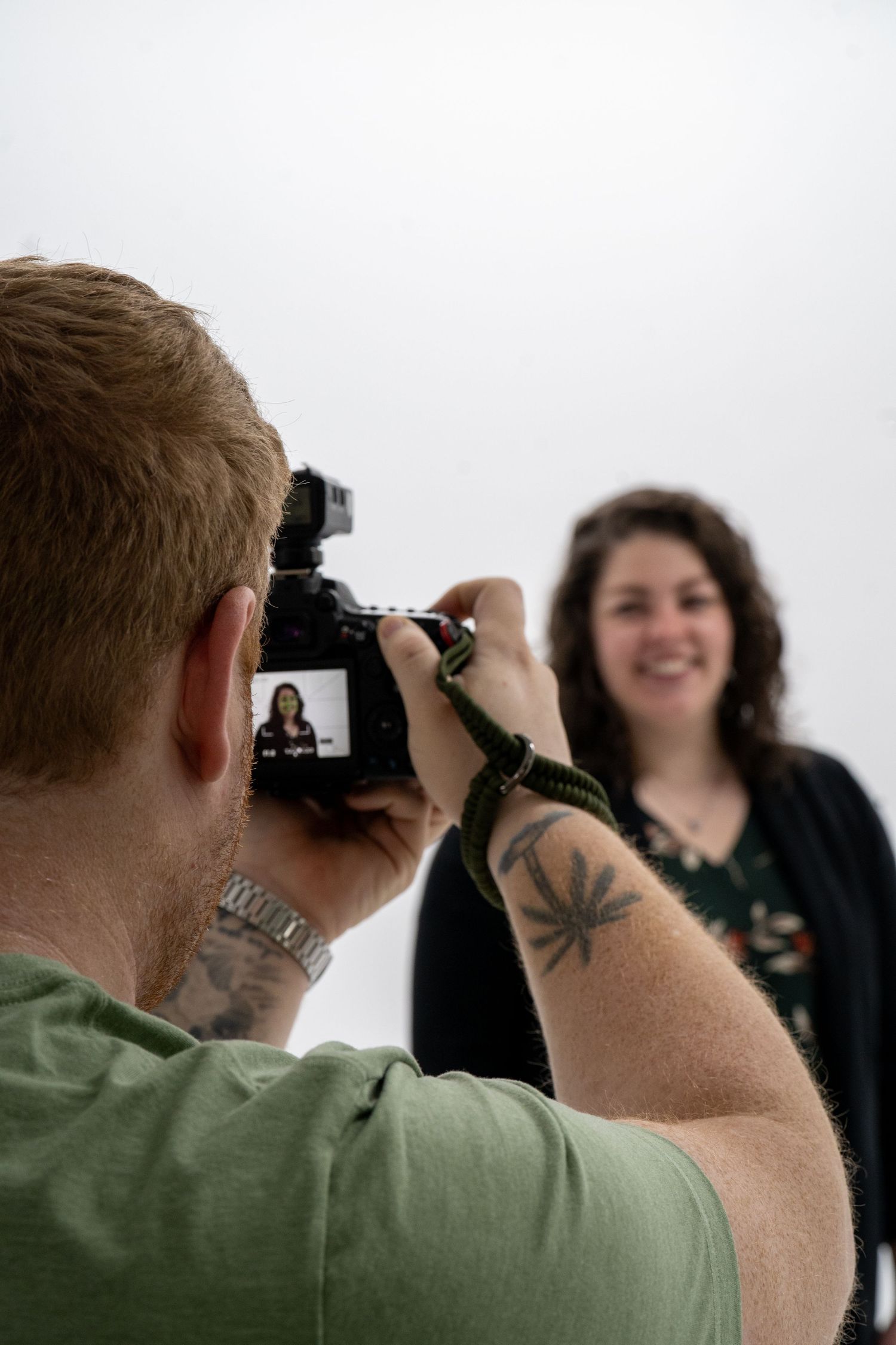 A photographer with a tattooed arm takes a photo of a subject in a studio against a white background.