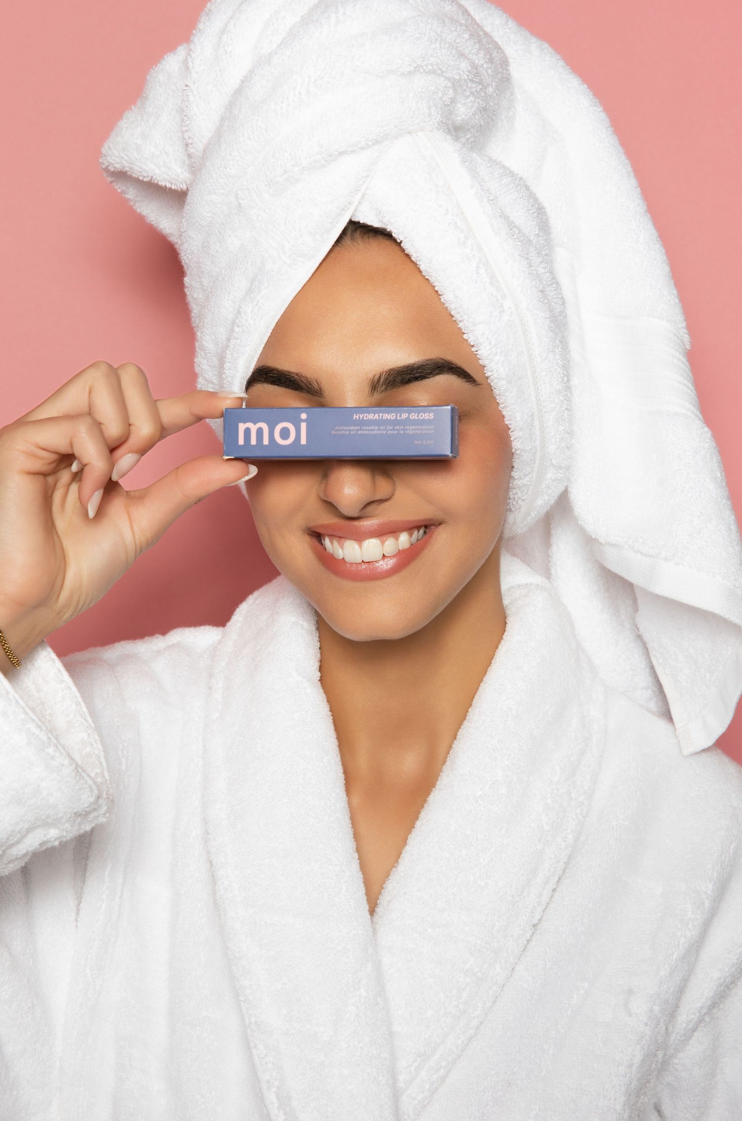 Woman in white bathrobe and towel turban smiling while holding a small blue Moi skincare product box.