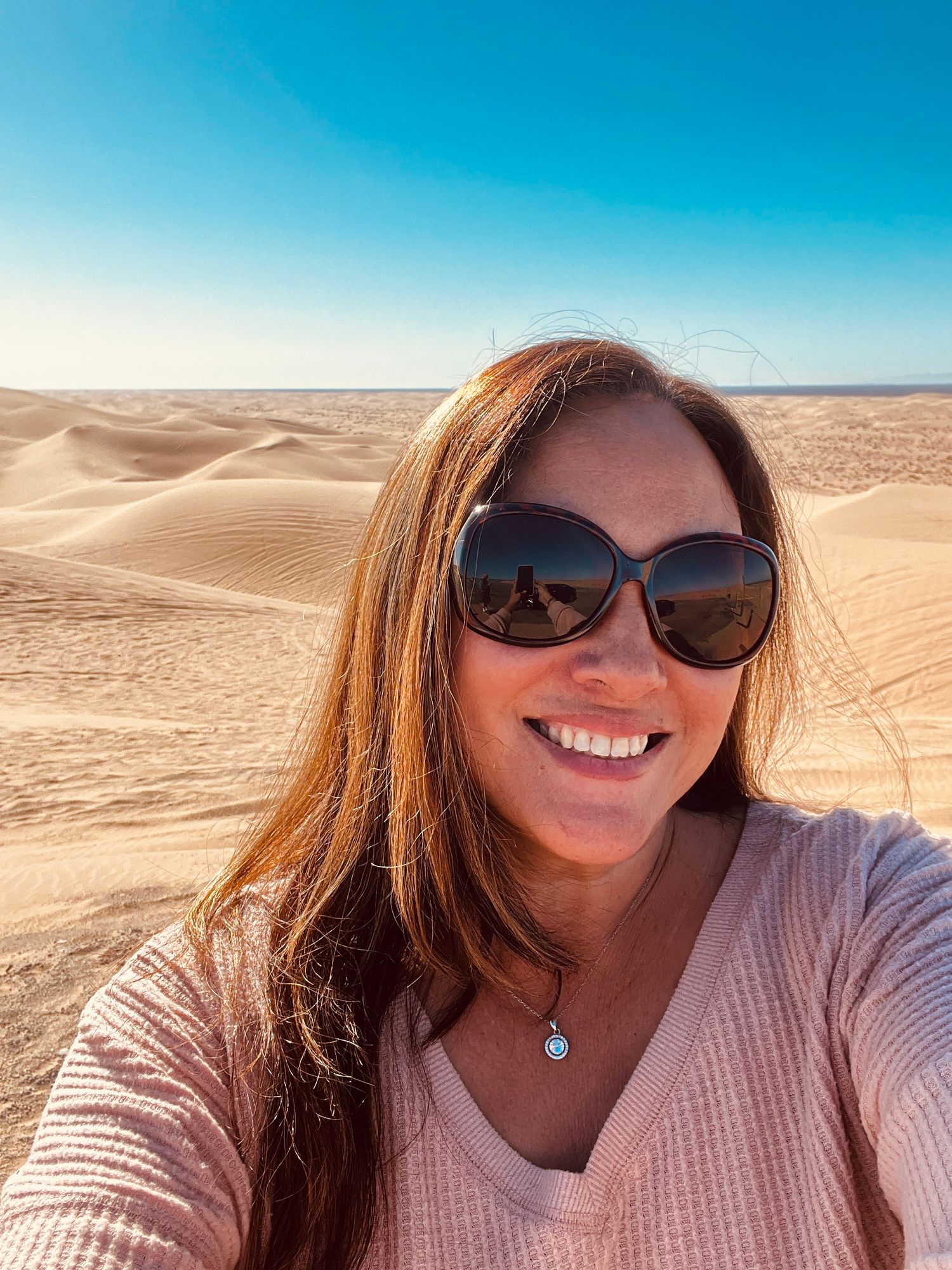 Woman smiling in desert selfie wearing sunglasses with sand dunes and blue sky background.