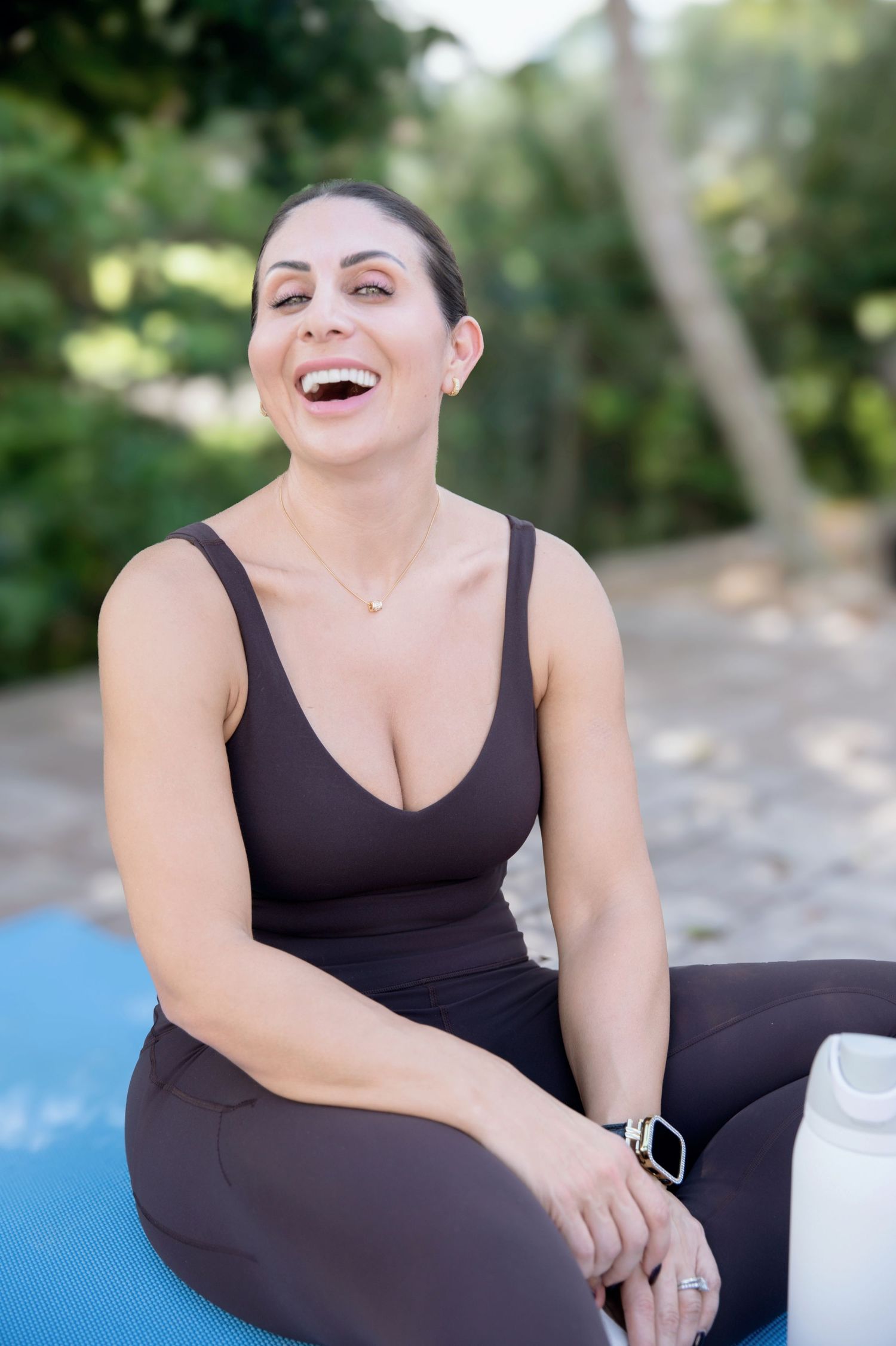Woman in dark athletic wear laughing outdoors on yoga mat with water bottle, enjoying fitness break in park.