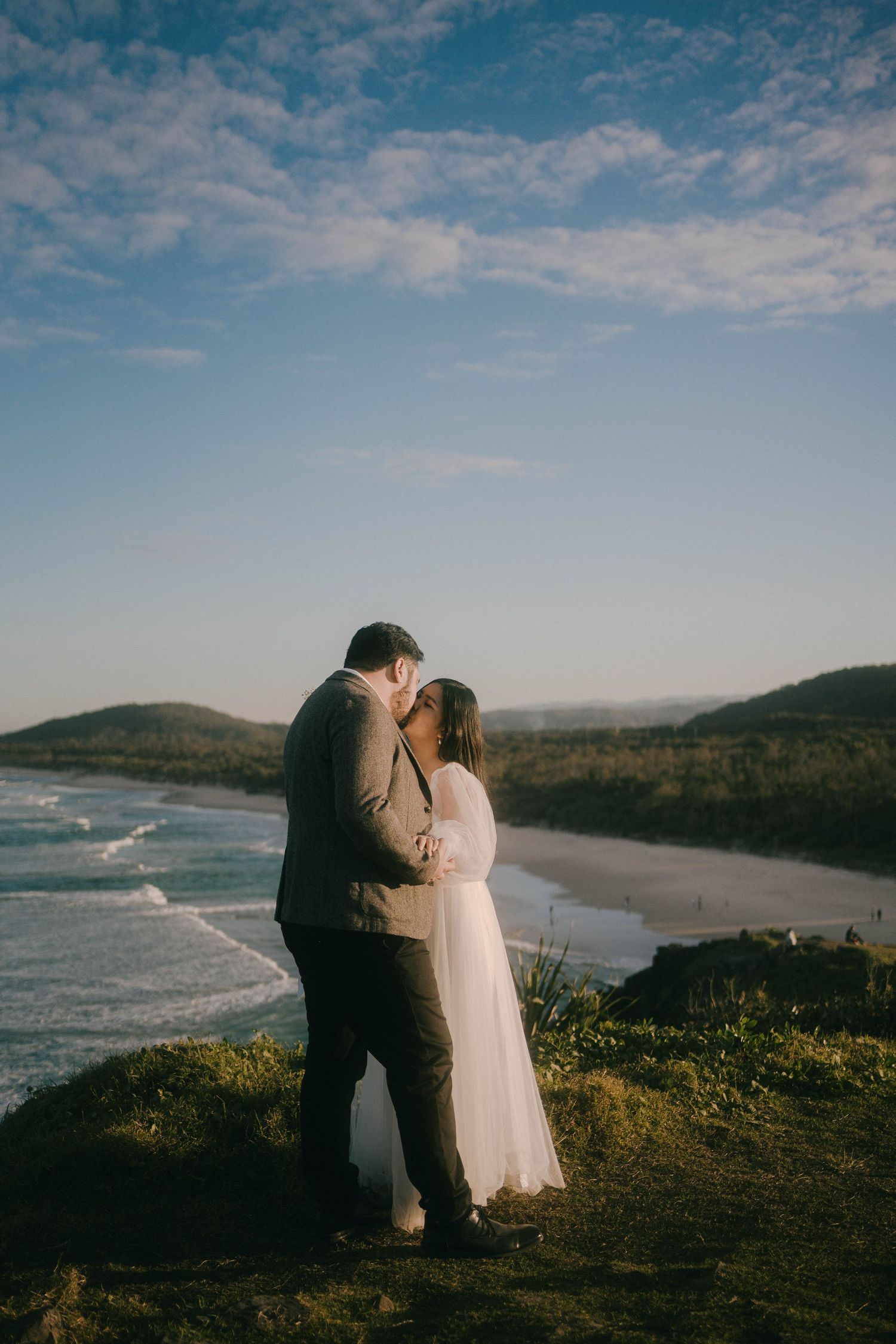 A couple embraces on a cliff overlooking a beautiful beach coastline during a dreamy photoshoot.