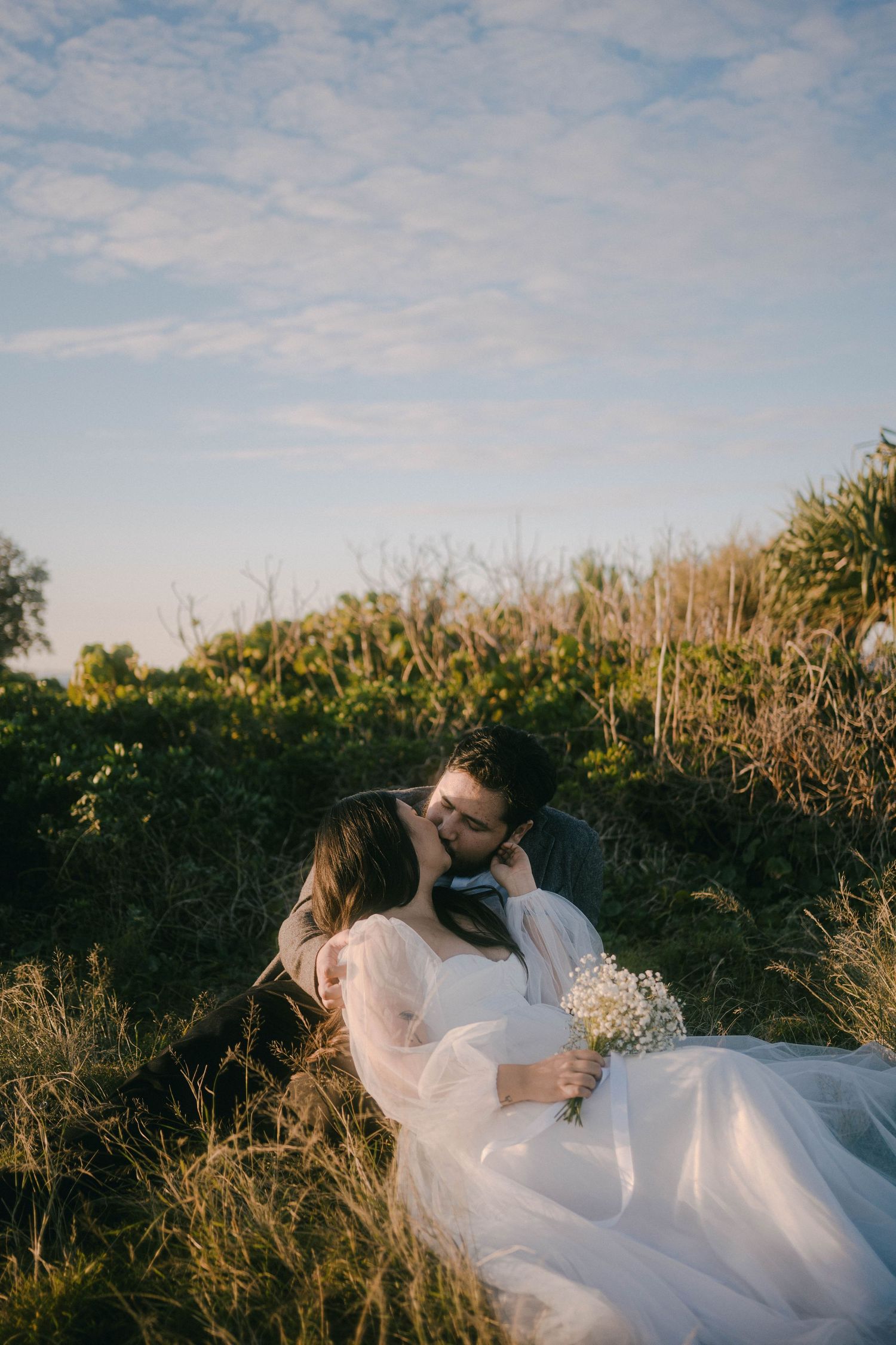 A romantic couple in a meadow at sunset, with the bride wearing a flowing white dress and holding baby's breath flowers.