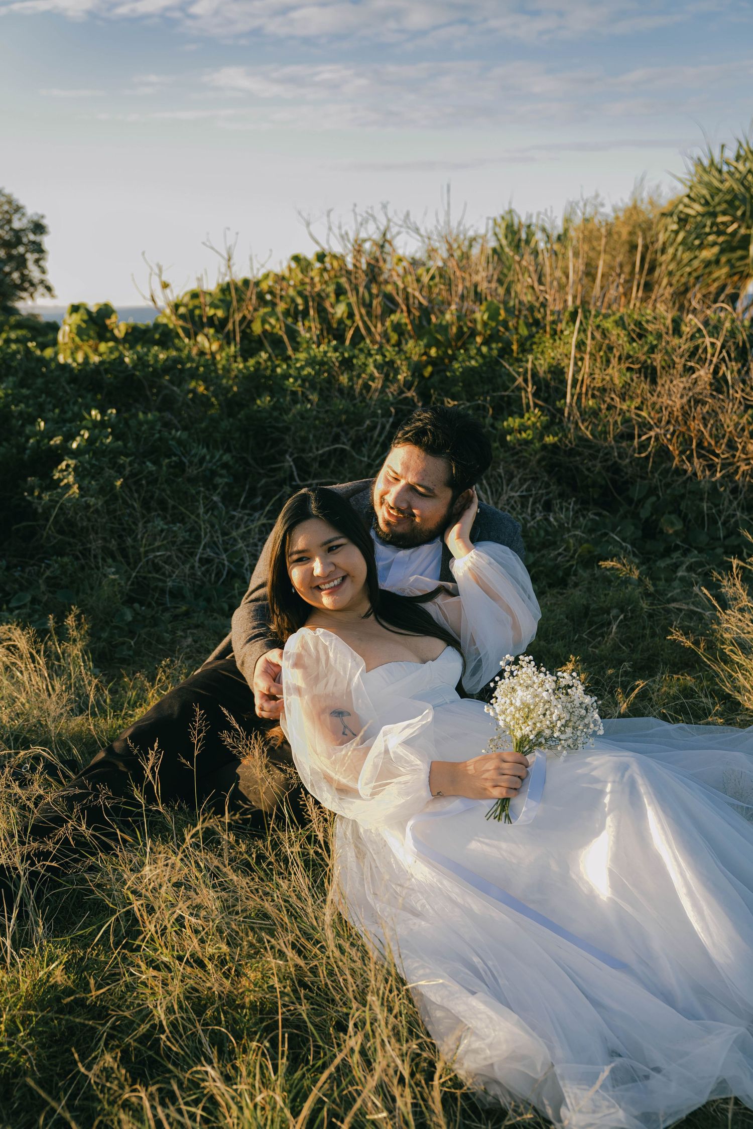 A couple in wedding attire embraces among tall grass at sunset, creating a romantic outdoor photoshoot series.