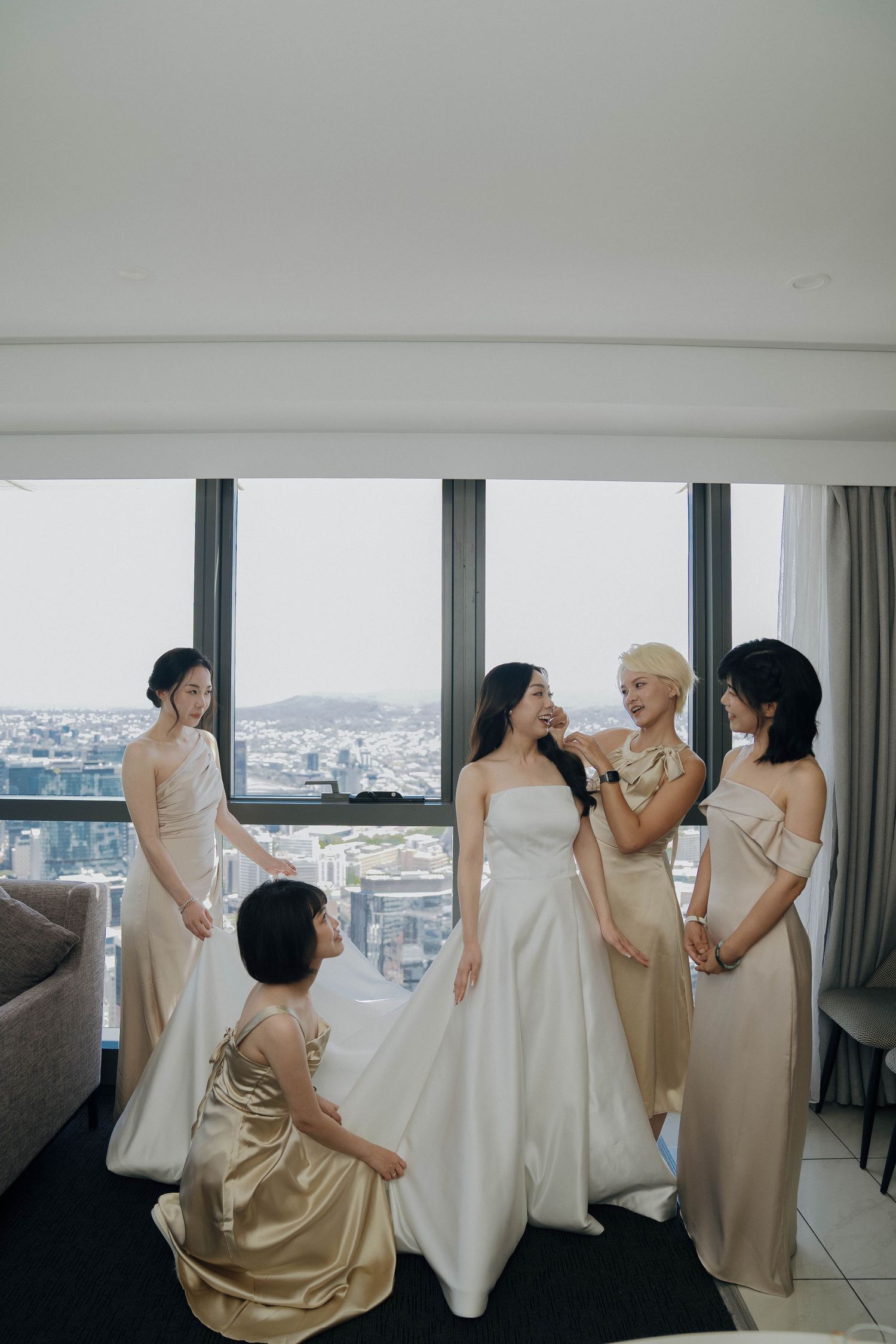 Bridesmaids in champagne dresses share joyful moments with the bride in a bright hotel room overlooking a city skyline.