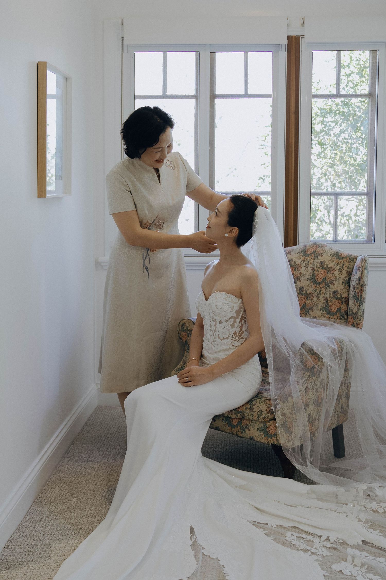 A wedding dress fitting session shows final adjustments being made in a bright sunlit room.