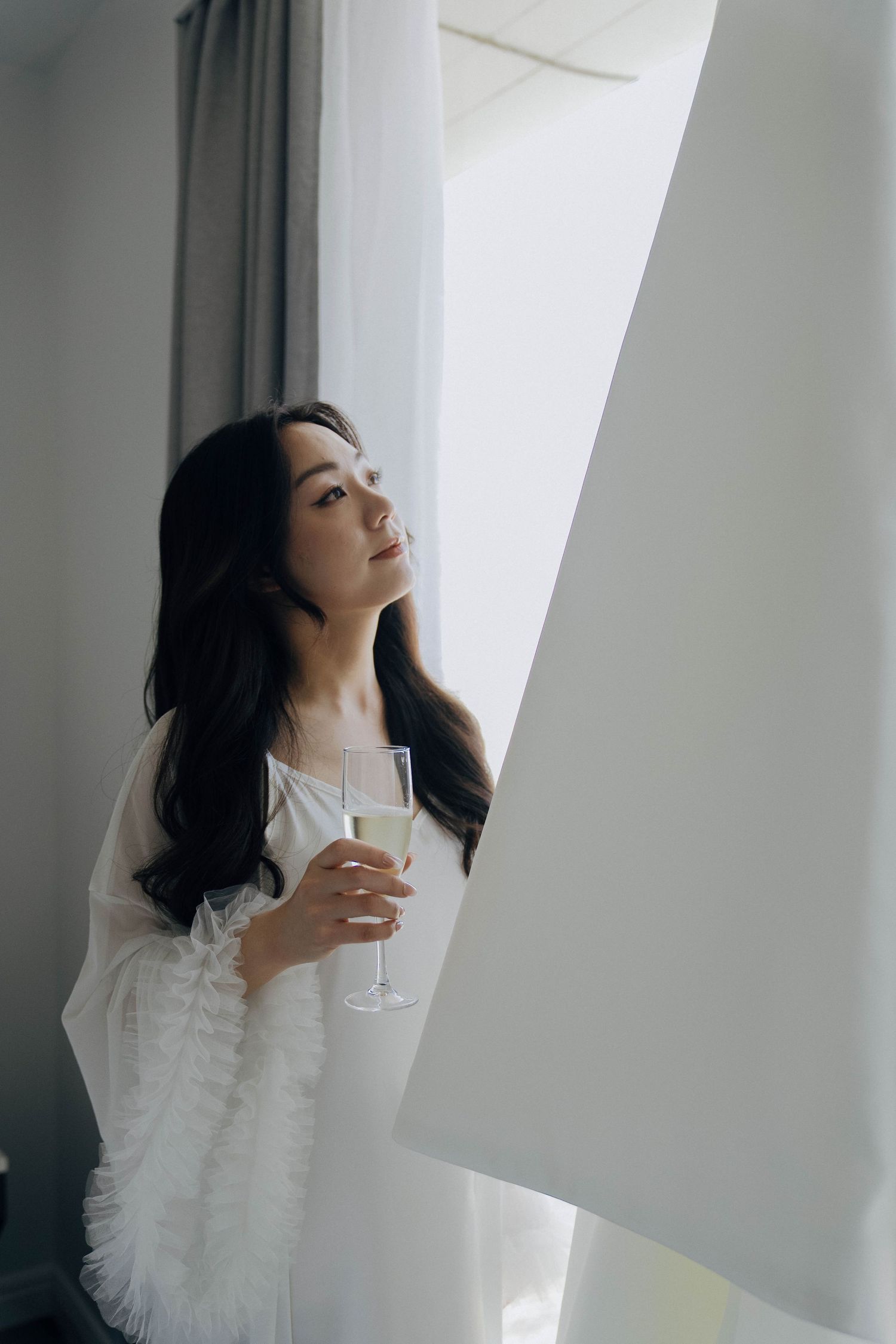 Person in white ruffled dress holding champagne glass by window with soft natural lighting.