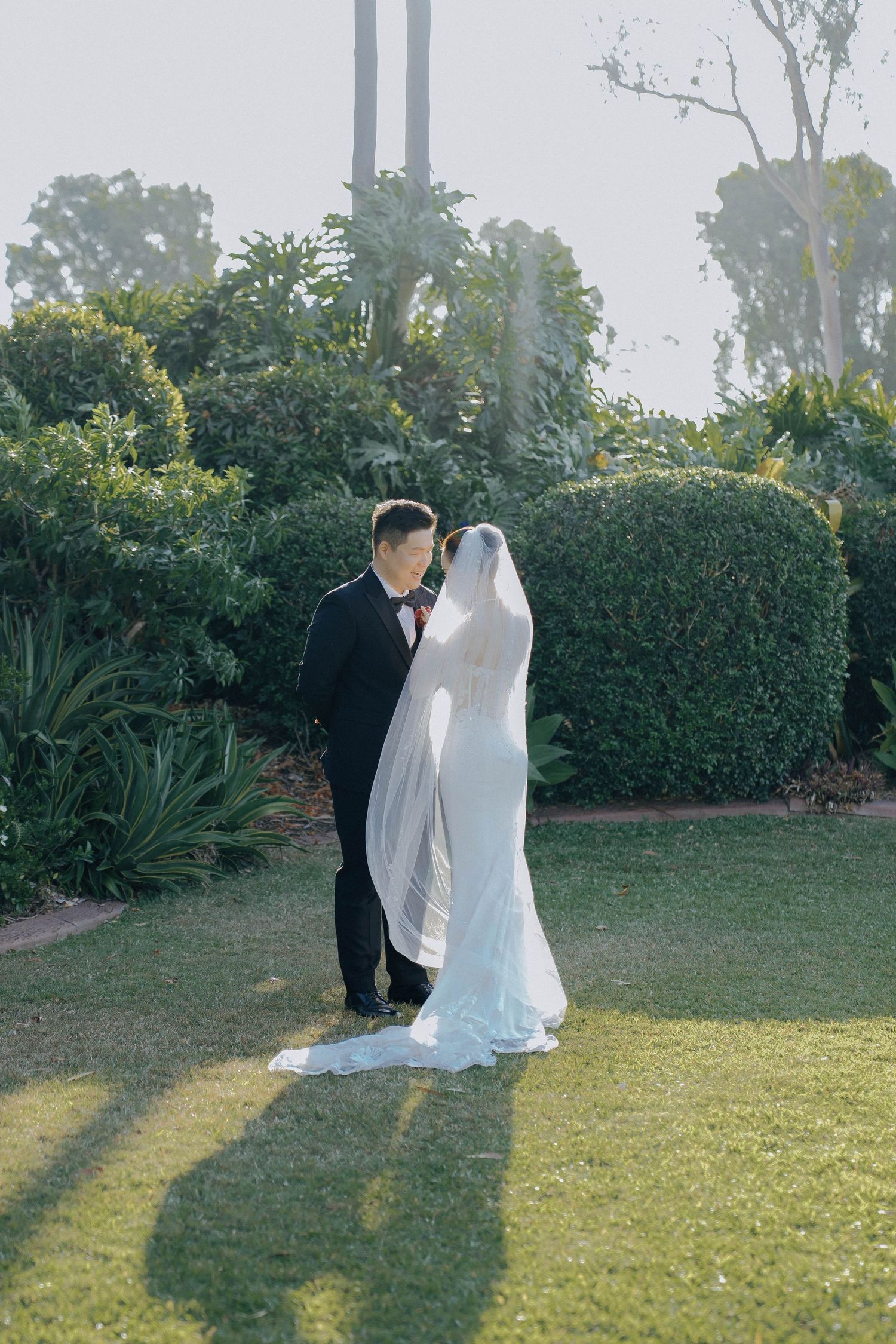 A couple in wedding attire share an intimate moment on a lawn with lush garden hedges and trees in the background.