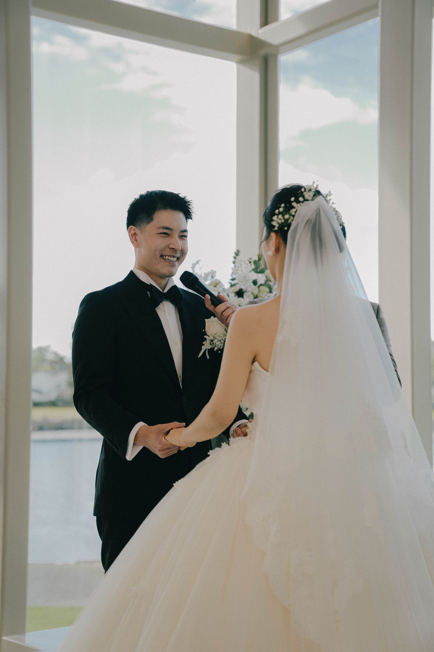 A couple exchanges vows in a bright ceremony space with floor-to-ceiling windows overlooking a scenic view.