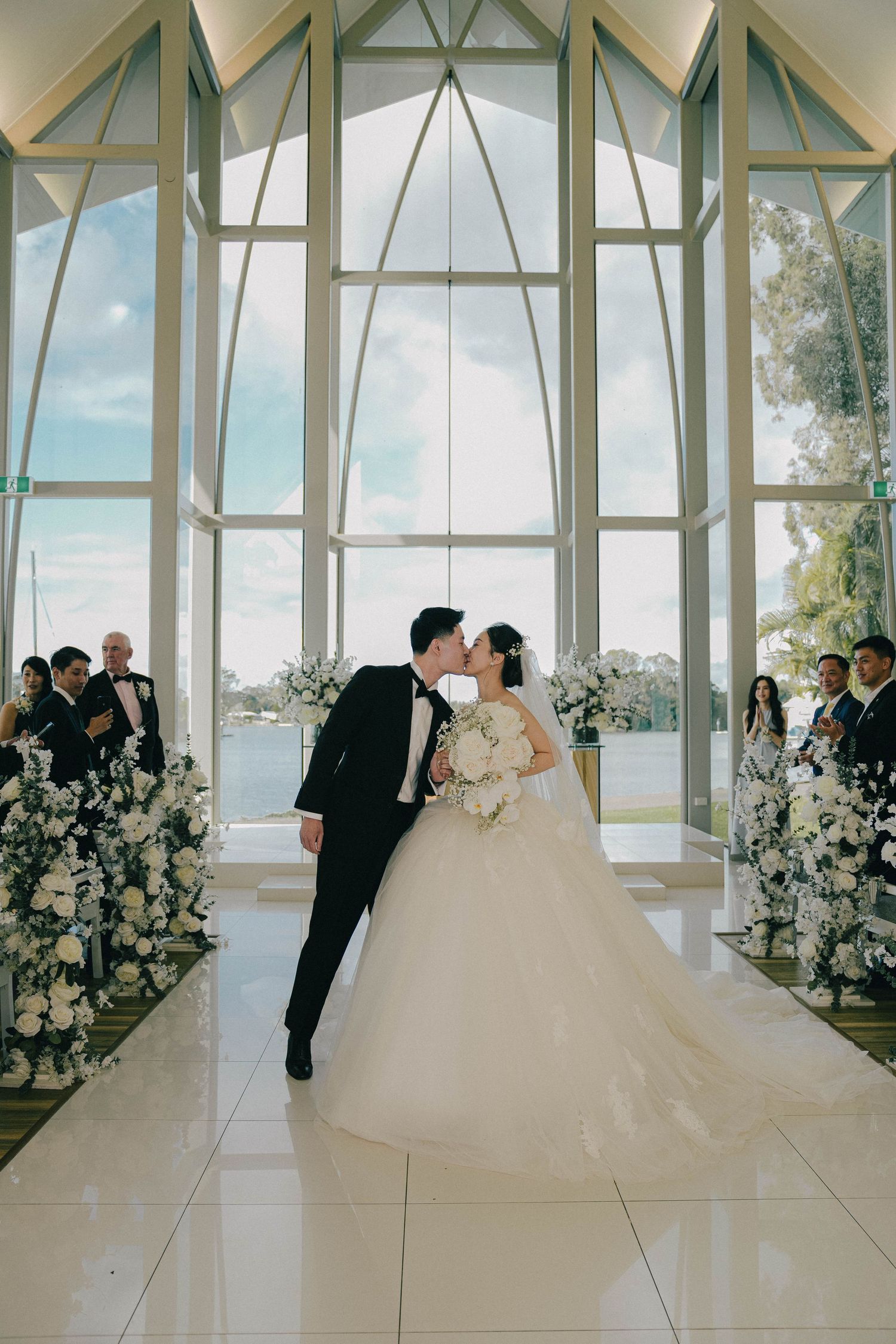 A couple shares a romantic moment in a modern chapel with floor-to-ceiling windows and elegant white floral arrangements.