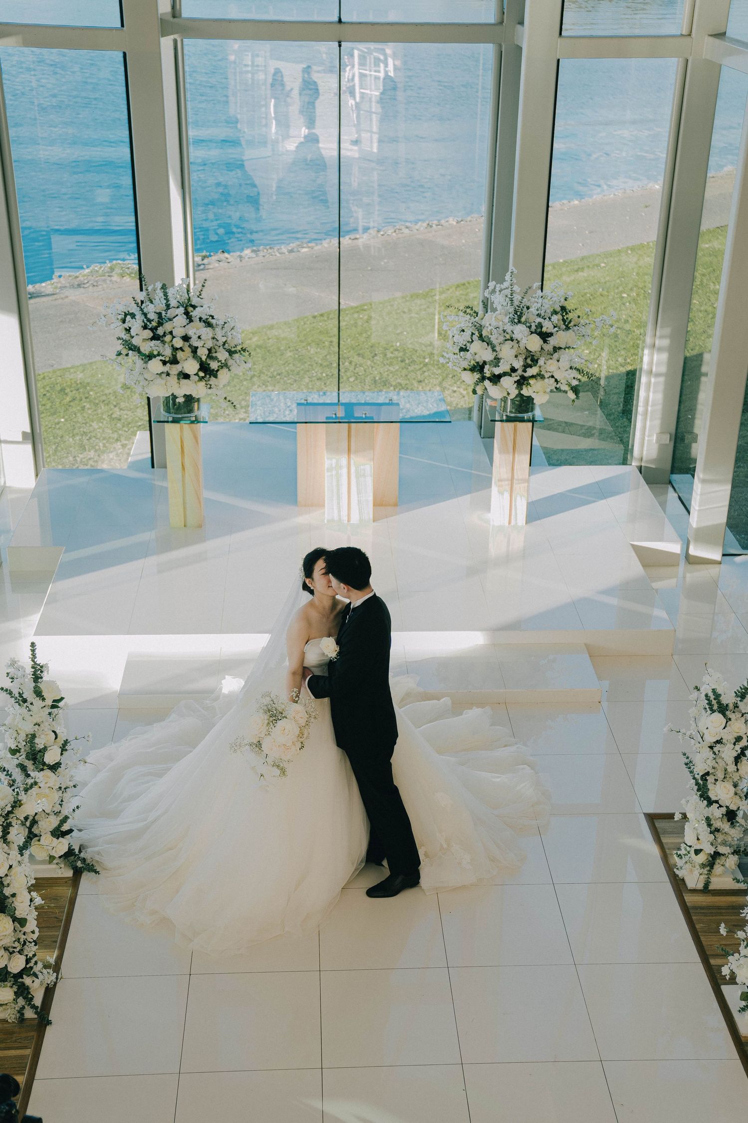 A wedding couple shares a romantic moment in a modern glass-walled venue overlooking the ocean.