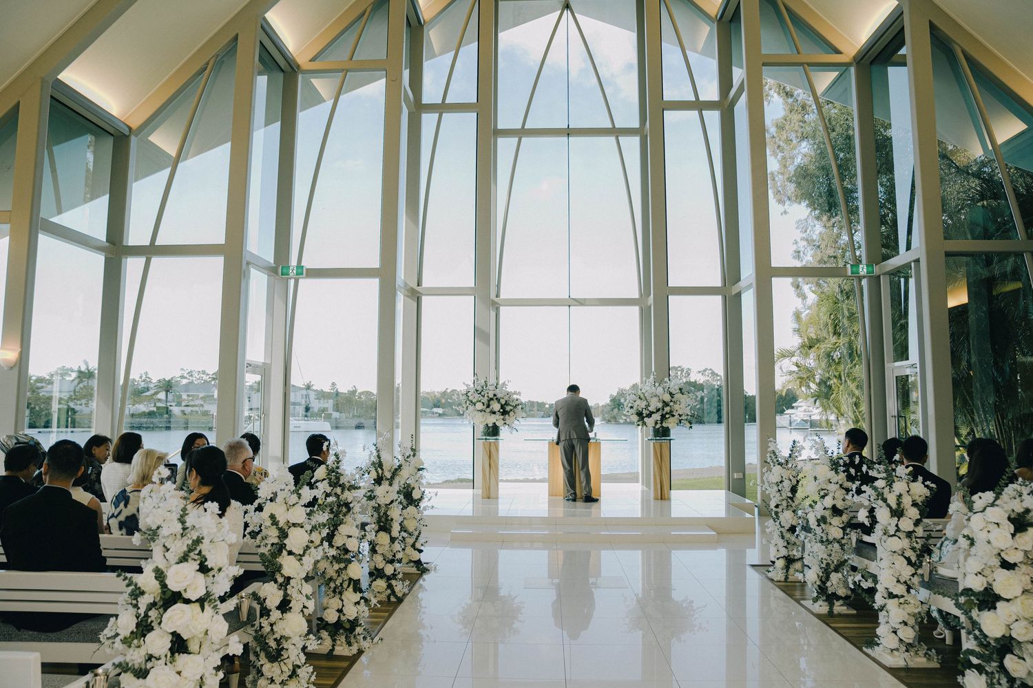 A modern glass-walled event space with white chairs and floral arrangements set up for a wedding ceremony.