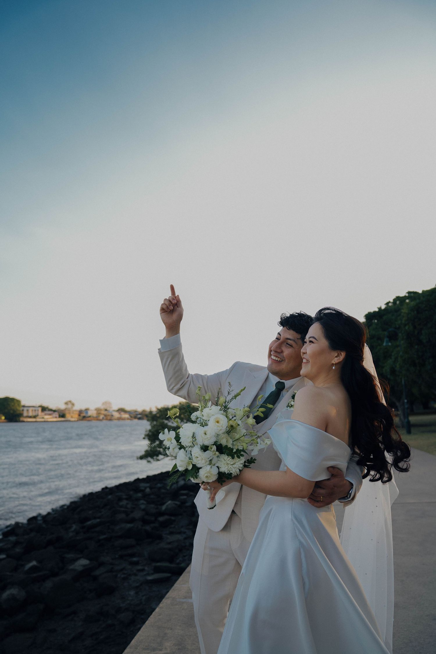 Newlywed couple embraces on rocky shoreline at sunset with white bouquet and ocean view in background.