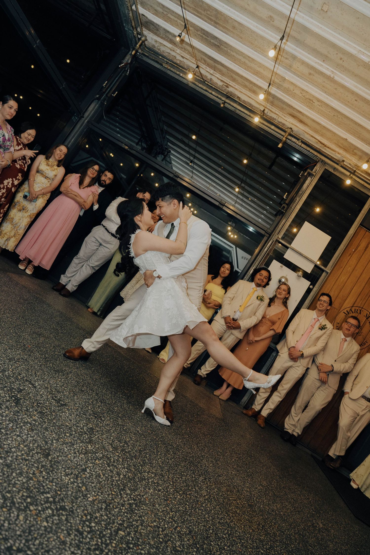 A wedding party poses together outside a venue at night with string lights glowing overhead.
