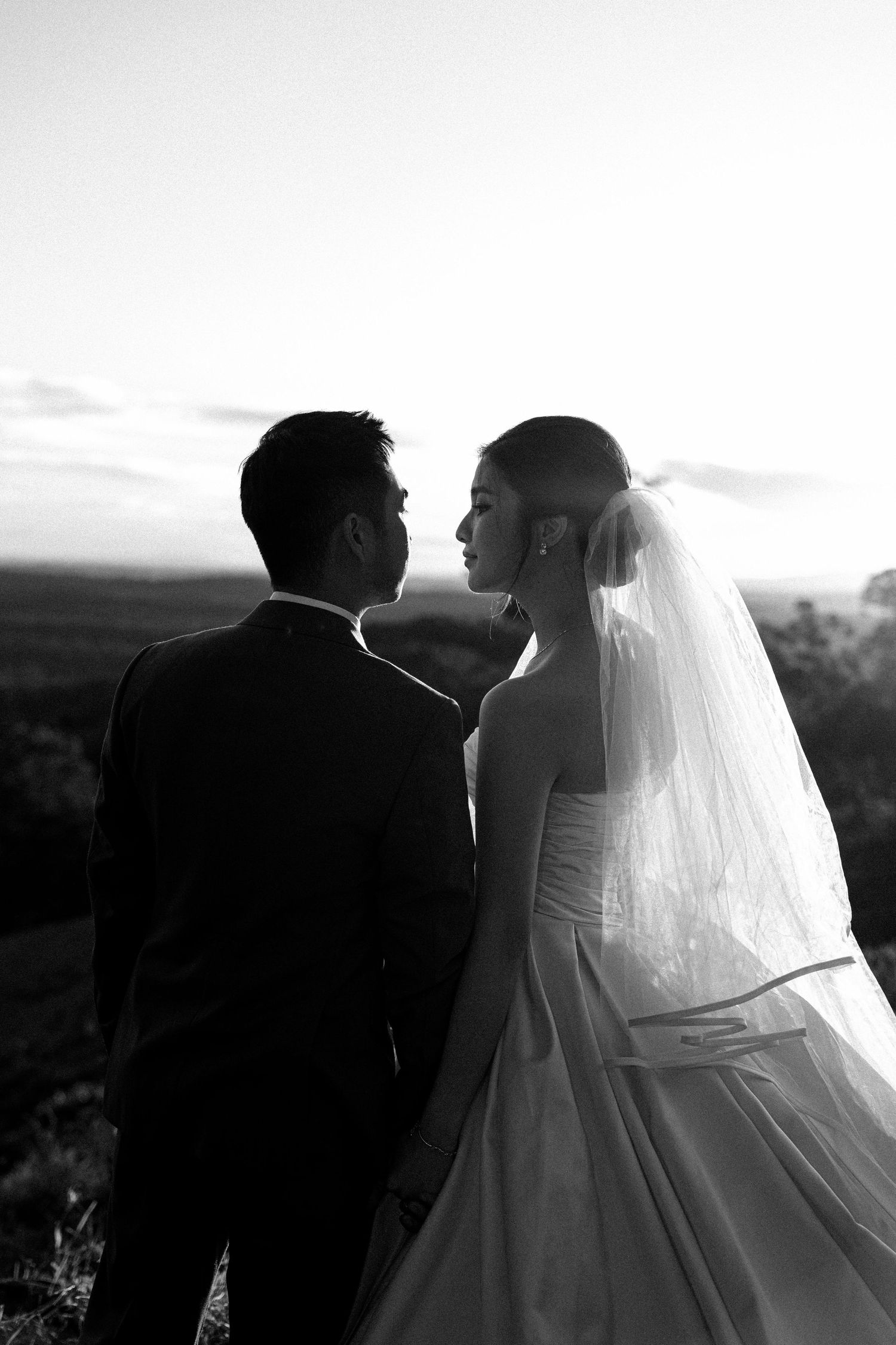 A silhouetted bride in a flowing veil and groom share a romantic moment against a sunset landscape.