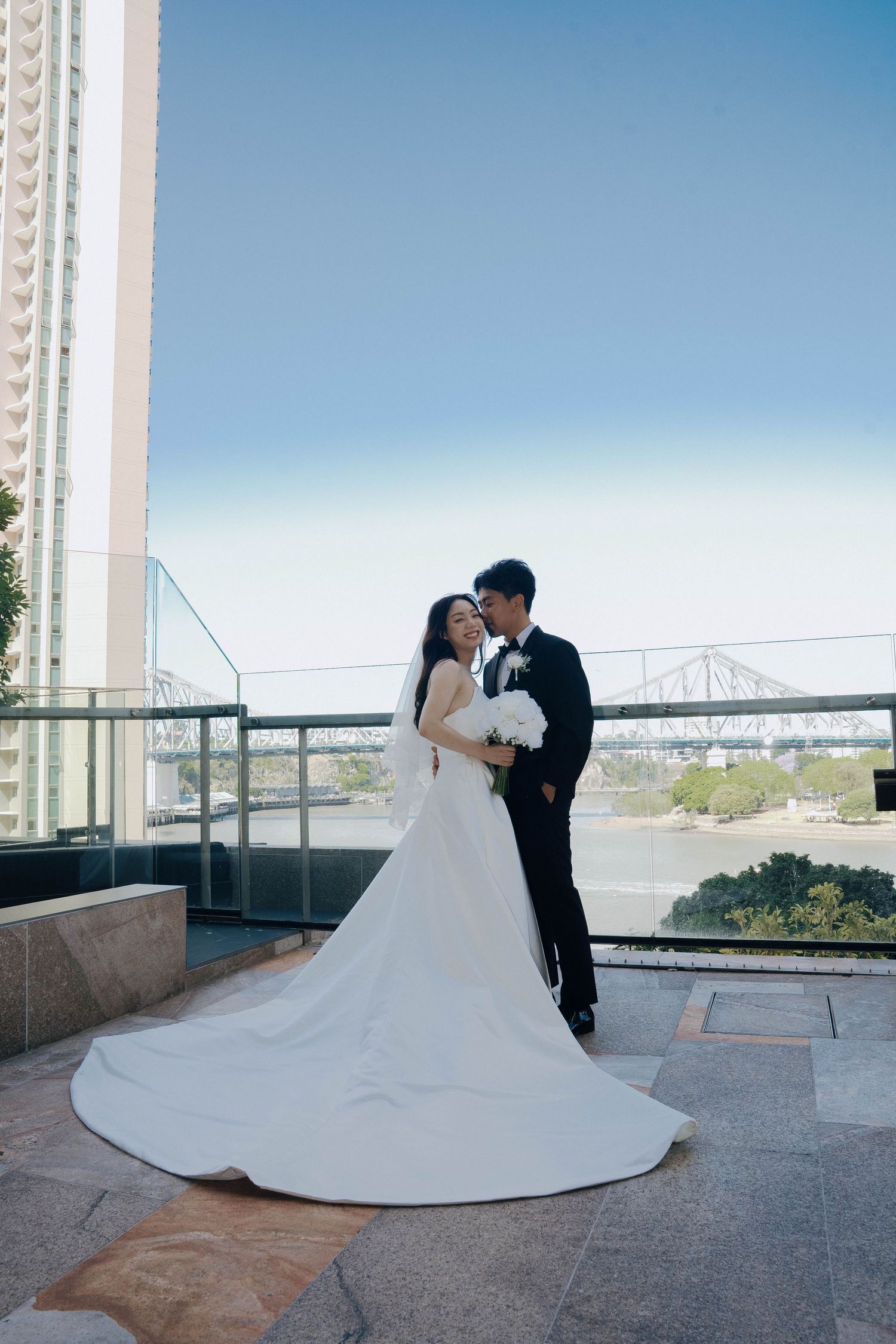A romantic couple in wedding attire share an intimate moment on a rooftop terrace with a bridge visible in the background.