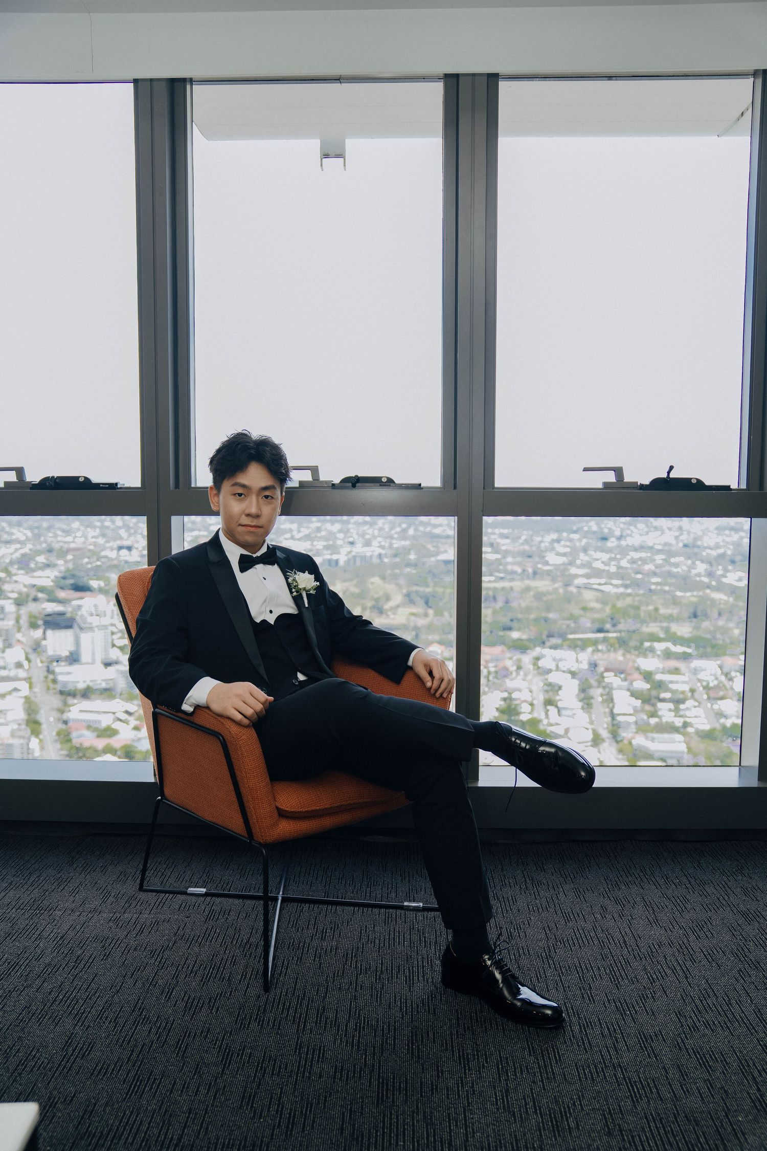 A person in black attire reclines on an orange chair against large windows overlooking a cityscape.