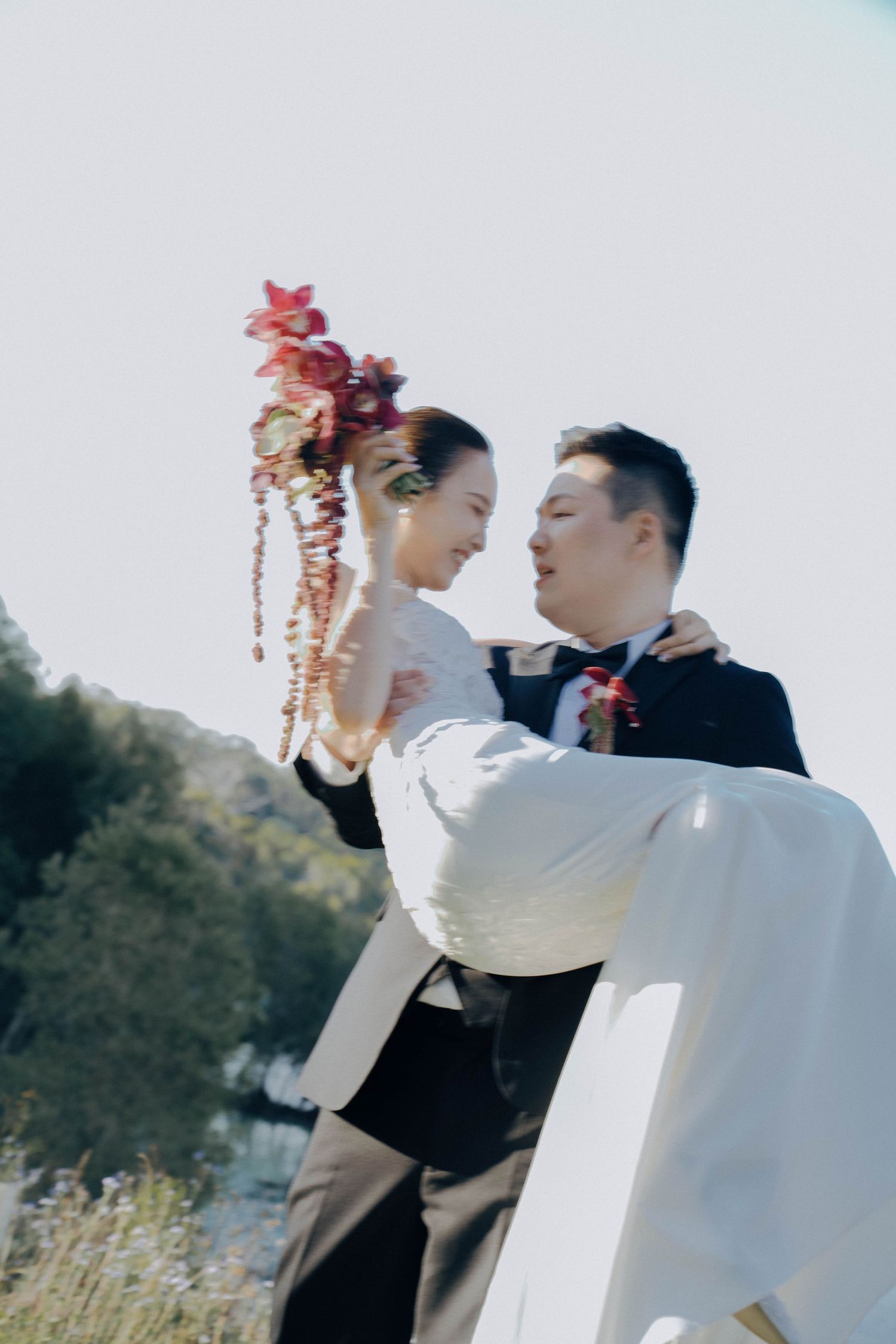 A romantic wedding couple embraces outdoors, with the bride wearing a white dress and groom in black suit.