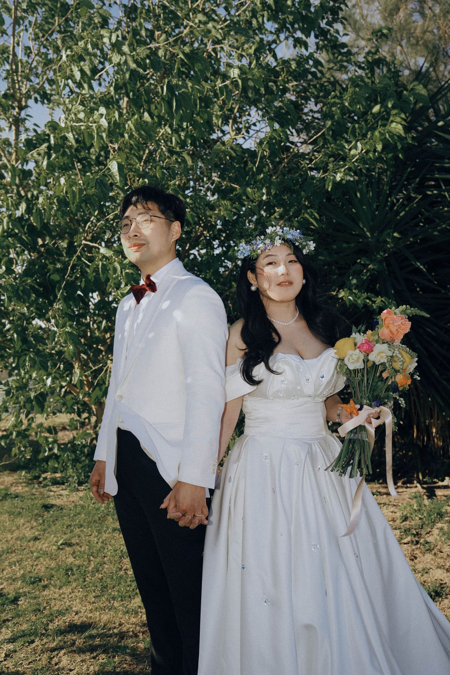 A couple in wedding attire stands together in a garden setting with the bride holding a colorful floral bouquet.
