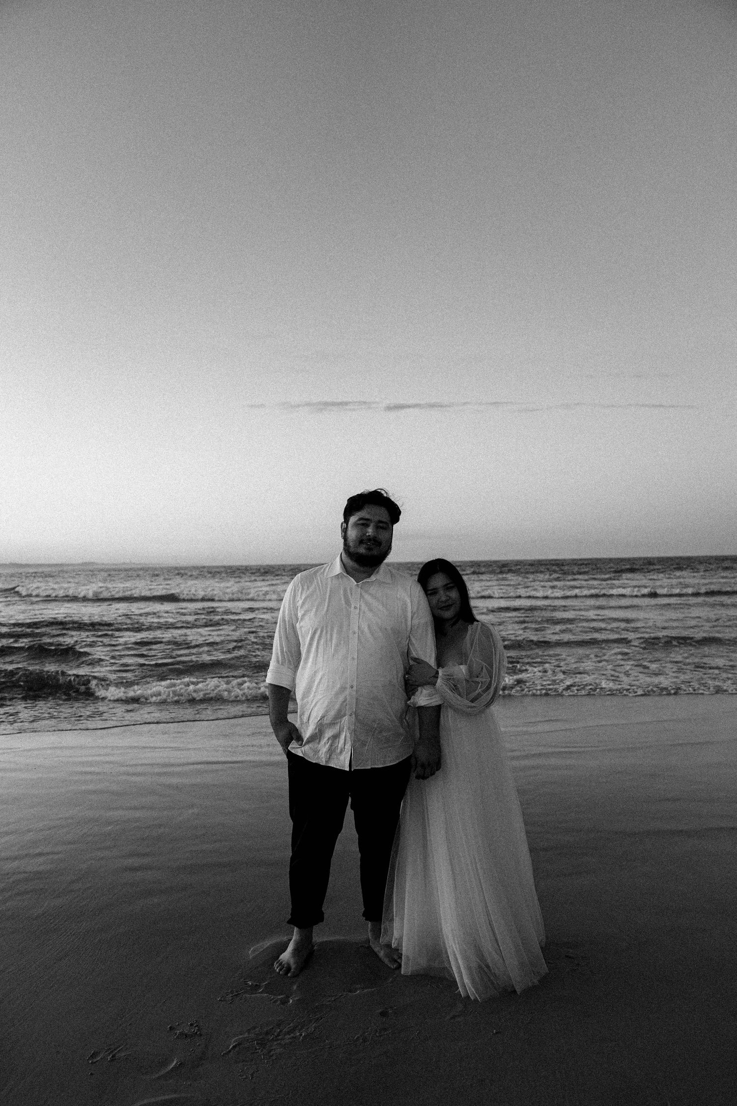 A couple stands together on a beach at sunset in a series of black and white photos showing their silhouettes against the ocean.