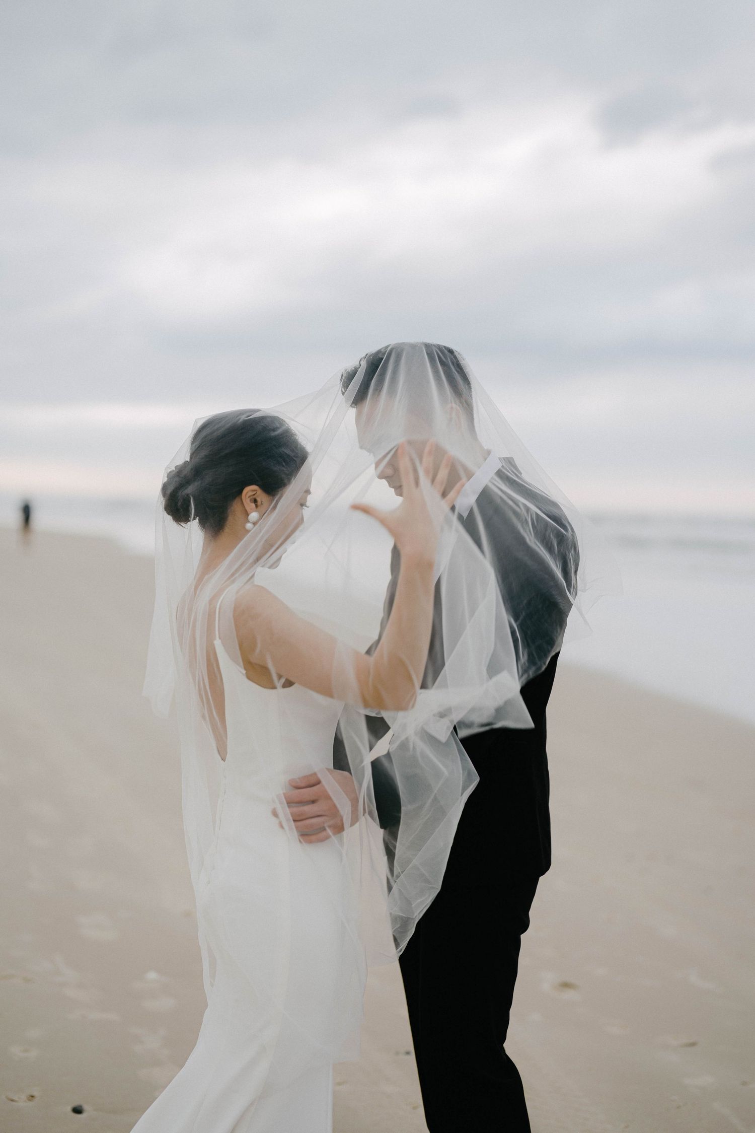 A romantic wedding couple embraces under a flowing veil on a misty beach with soft overcast lighting.