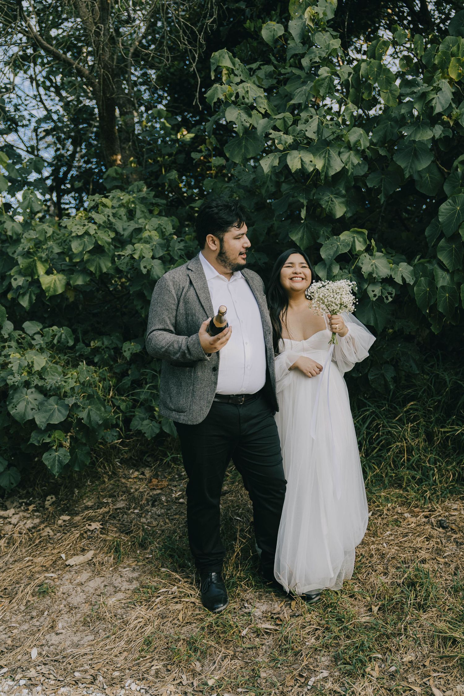 A couple in wedding attire stands together outdoors against lush green foliage on their special day.