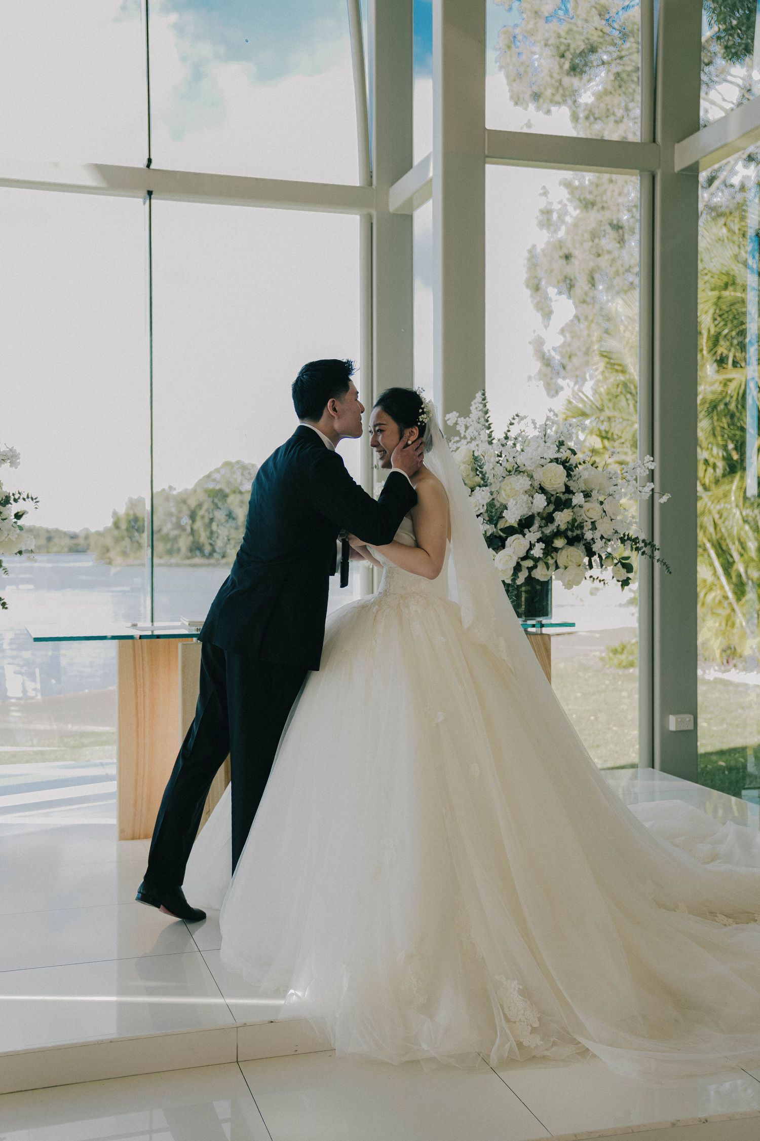 A romantic wedding moment captured in a bright sunlit chapel with floor-to-ceiling windows and white floral arrangements.