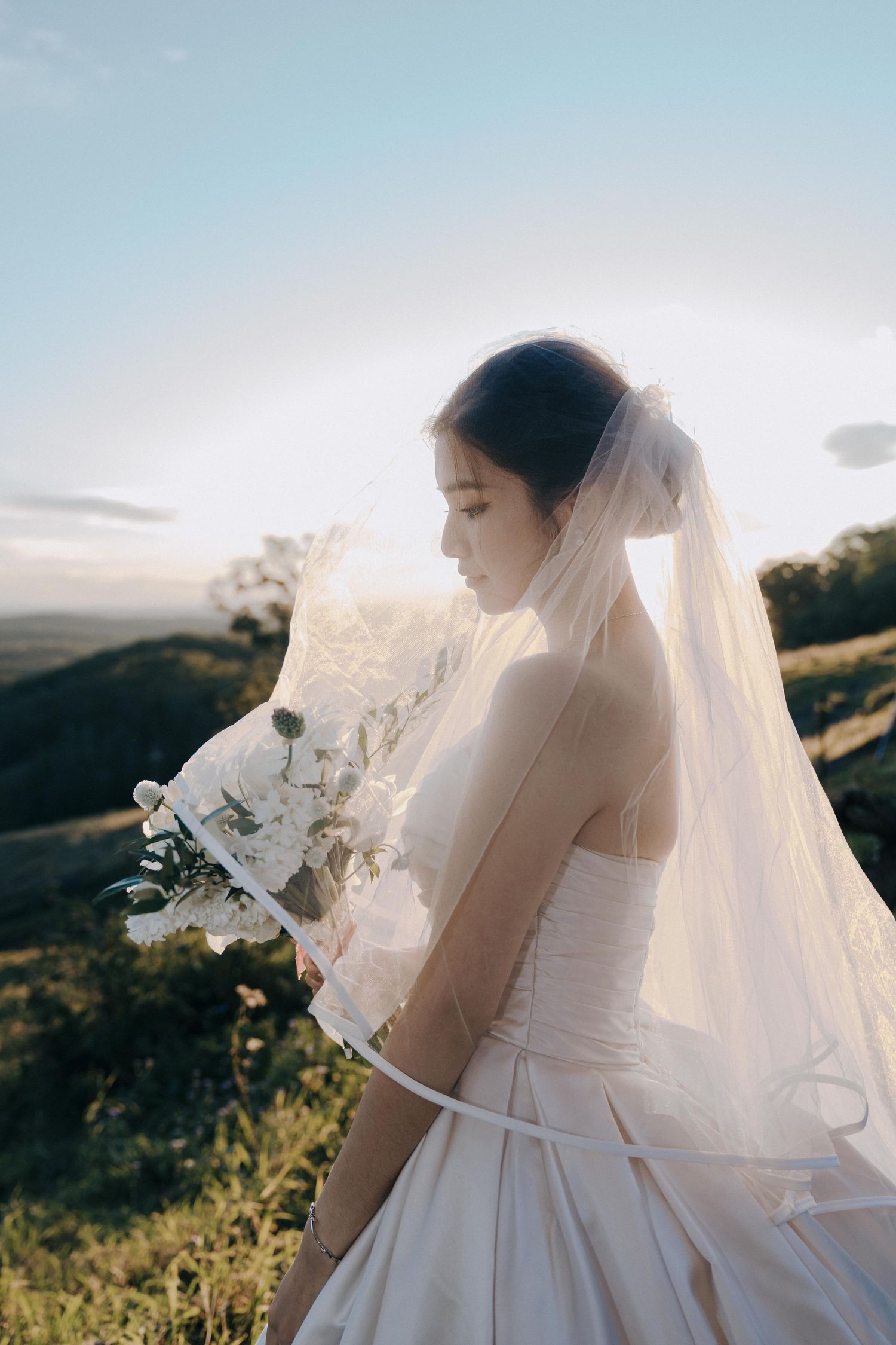 A bride in a classic white wedding dress stands outdoors at sunset with a flowing veil in the warm evening light.