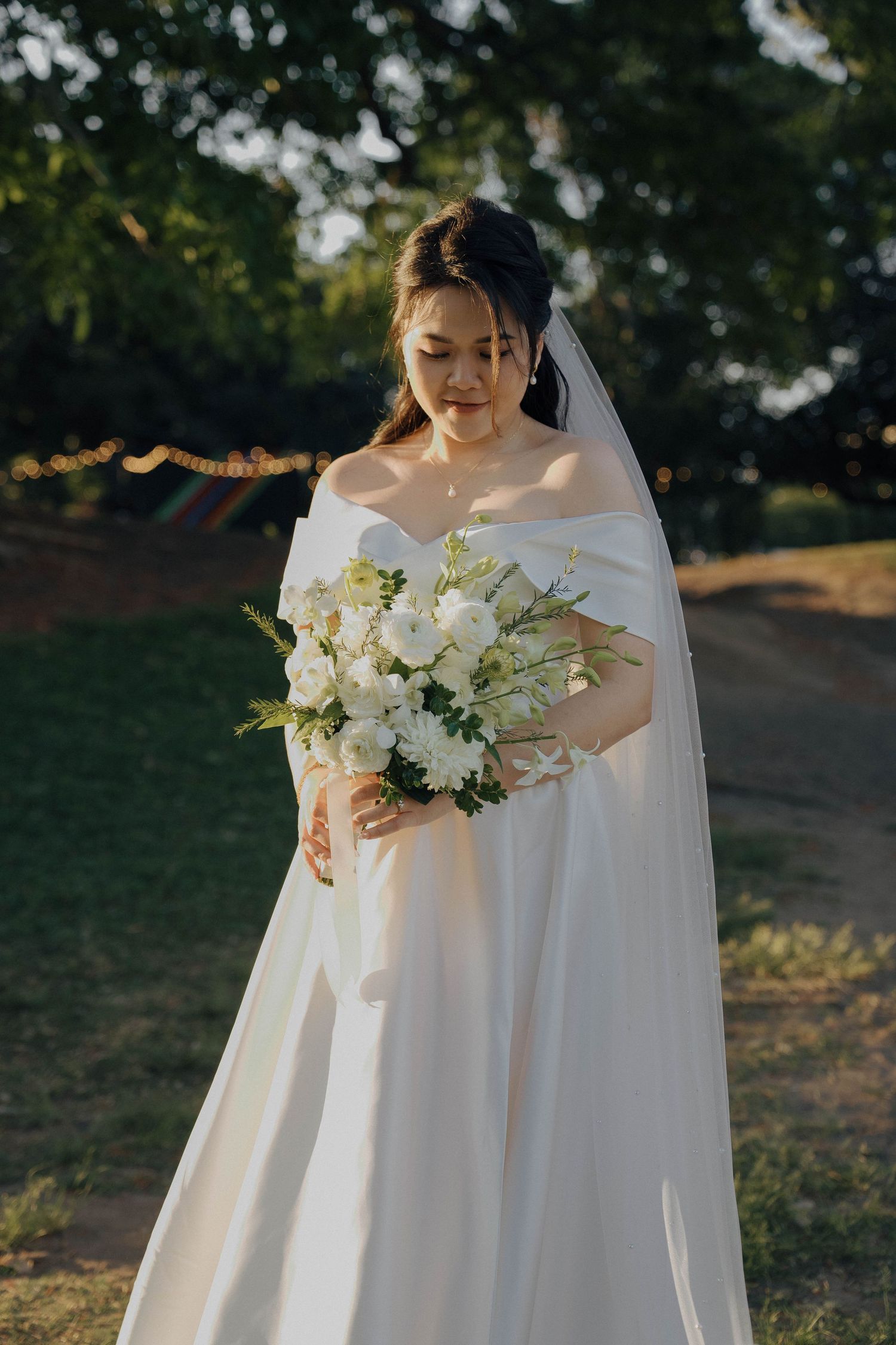 A bride in an elegant white off-shoulder gown holds a stunning bouquet of white flowers in an outdoor garden setting.