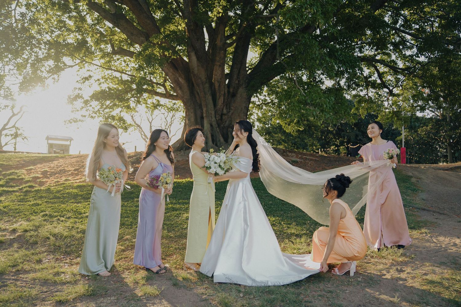 A group in pastel bridesmaid dresses and white wedding gown gather under sunlit trees in a park setting.